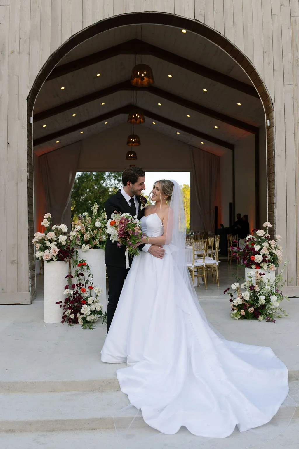 A bride and groom standing together outside a wedding venue, with floral arrangements around them, smiling at each other.