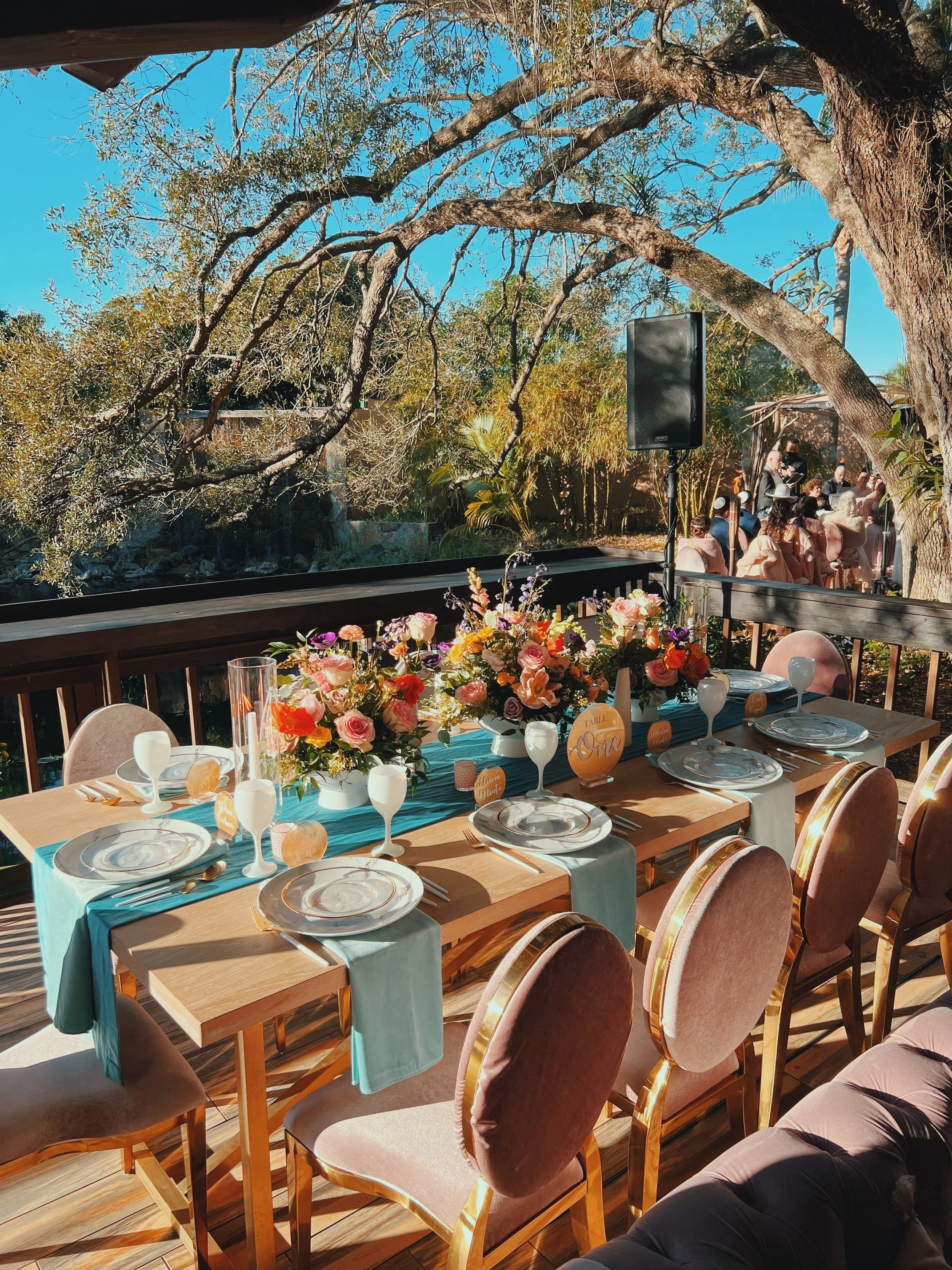 A decorated outdoor dining table with floral centerpieces, place settings, and glasses, set on a wooden deck under a large tree, with a gathering of people in the background.