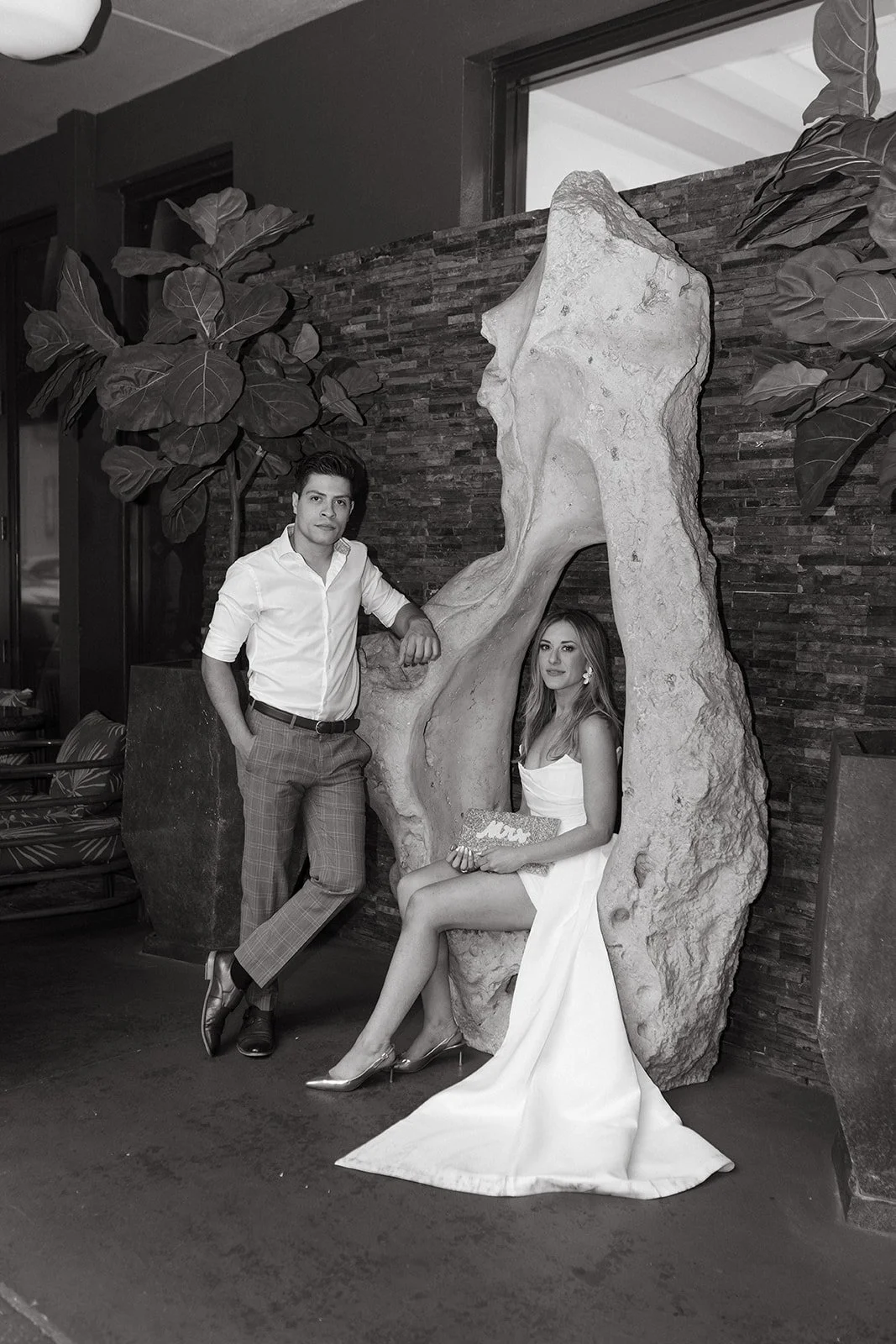 A man and woman in formal attire posing indoors. The woman is sitting on the floor wearing a white gown, holding a clutch, with her legs crossed. The man is standing beside her, leaning on a large sculptural rock, dressed in a white shirt and plaid p