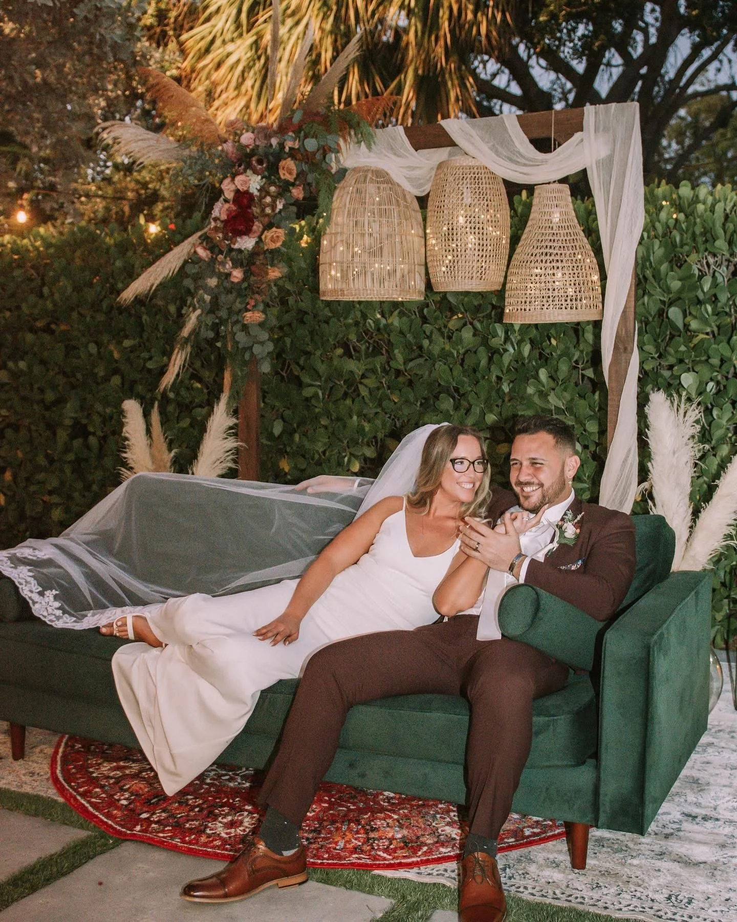 A bride and groom are sitting together on a green velvet sofa under a decorated outdoor wedding arch with hanging wicker lanterns, surrounded by greenery and flowers, during sunset.
