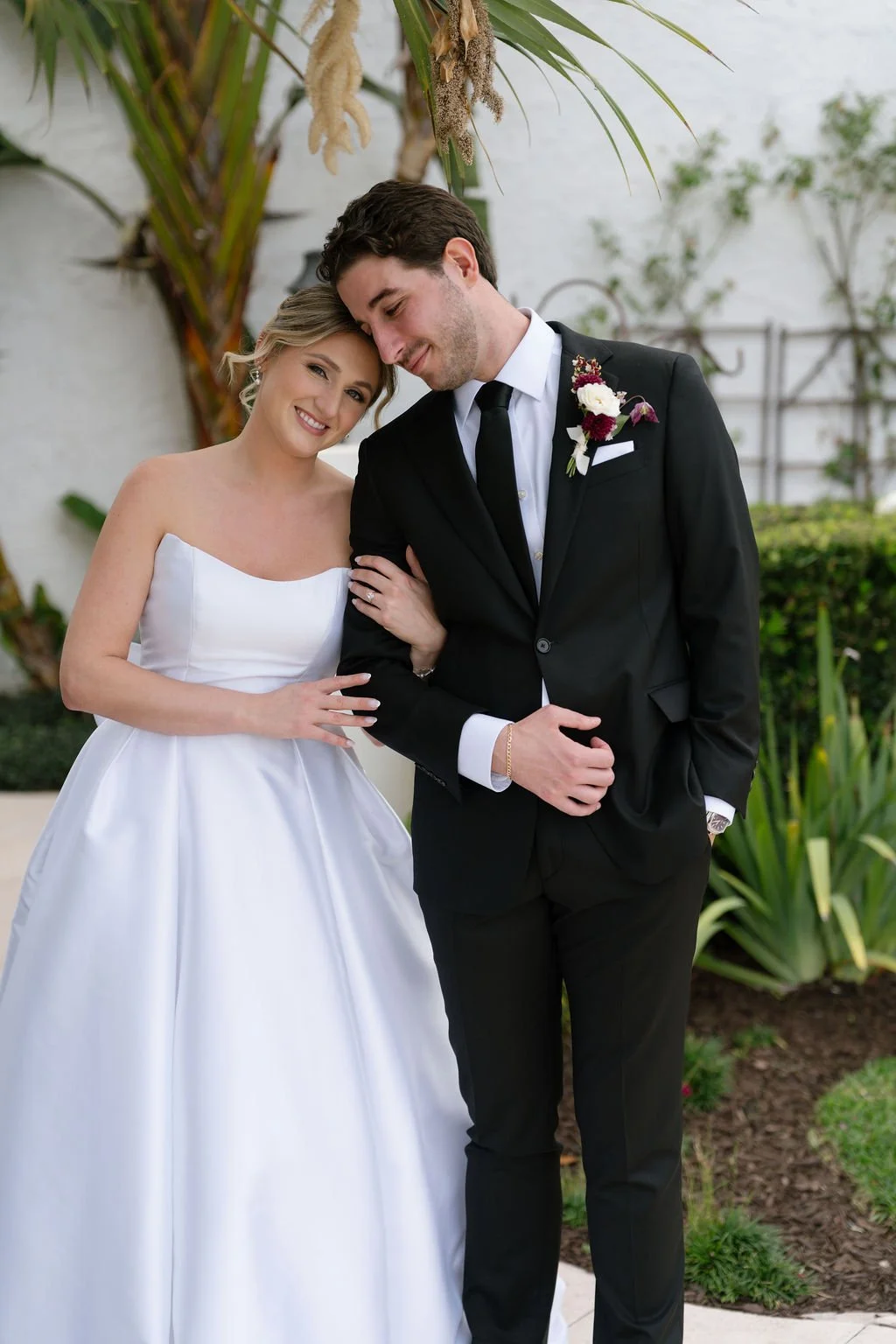 A bride and groom on their wedding day, smiling and leaning their heads together, outdoors with lush greenery and palm trees in the background.