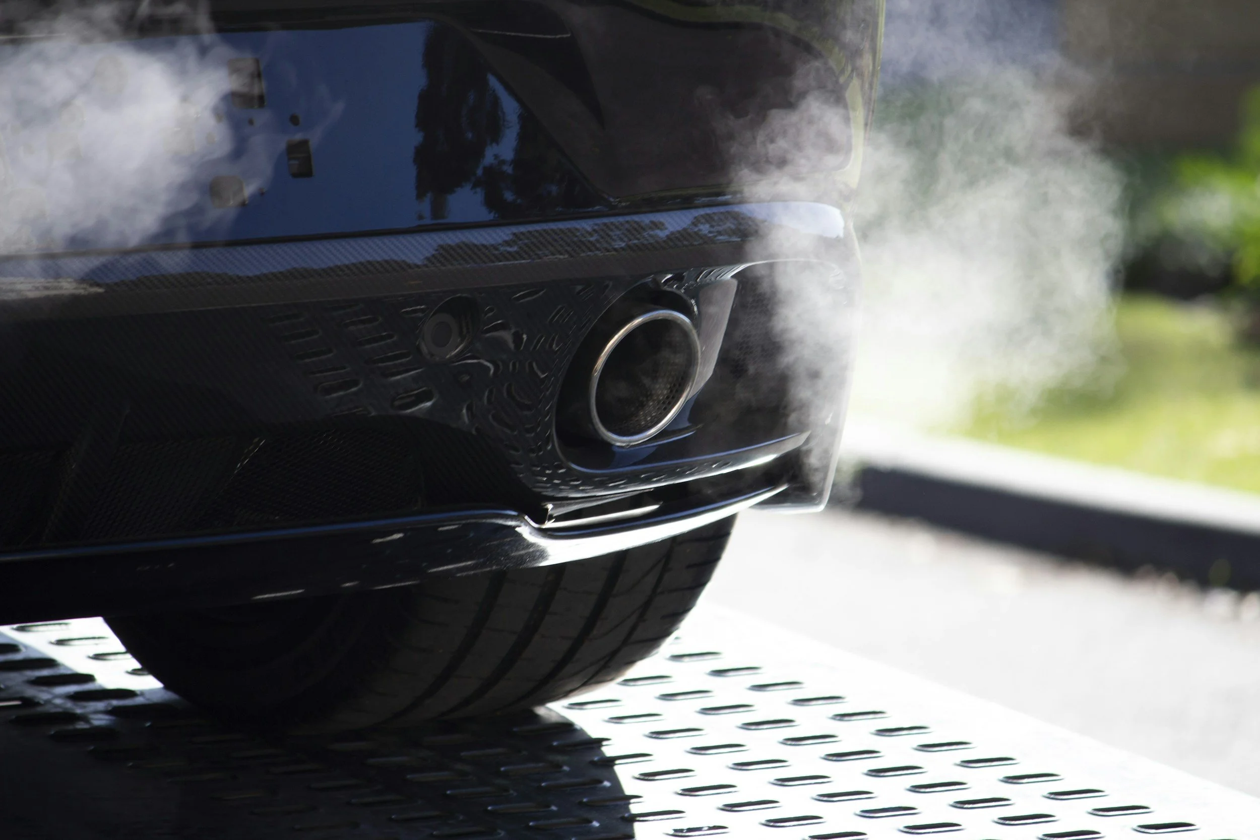 A black sports car's rear end with visible exhaust pipe emitting white smoke or vapor, on a grated metal surface.