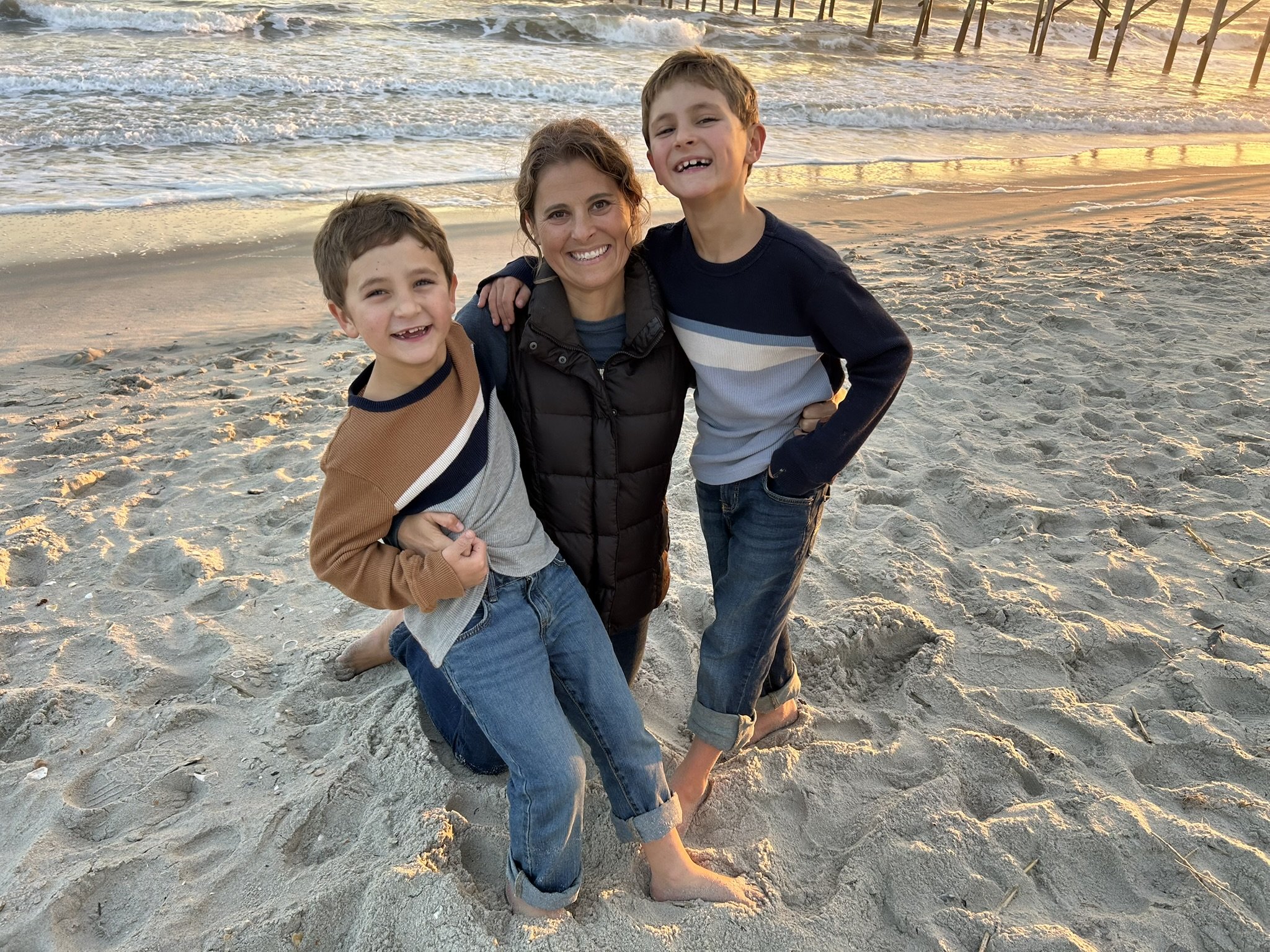A woman with two young boys smiling on a sandy beach during sunset, with waves and a pier in the background.