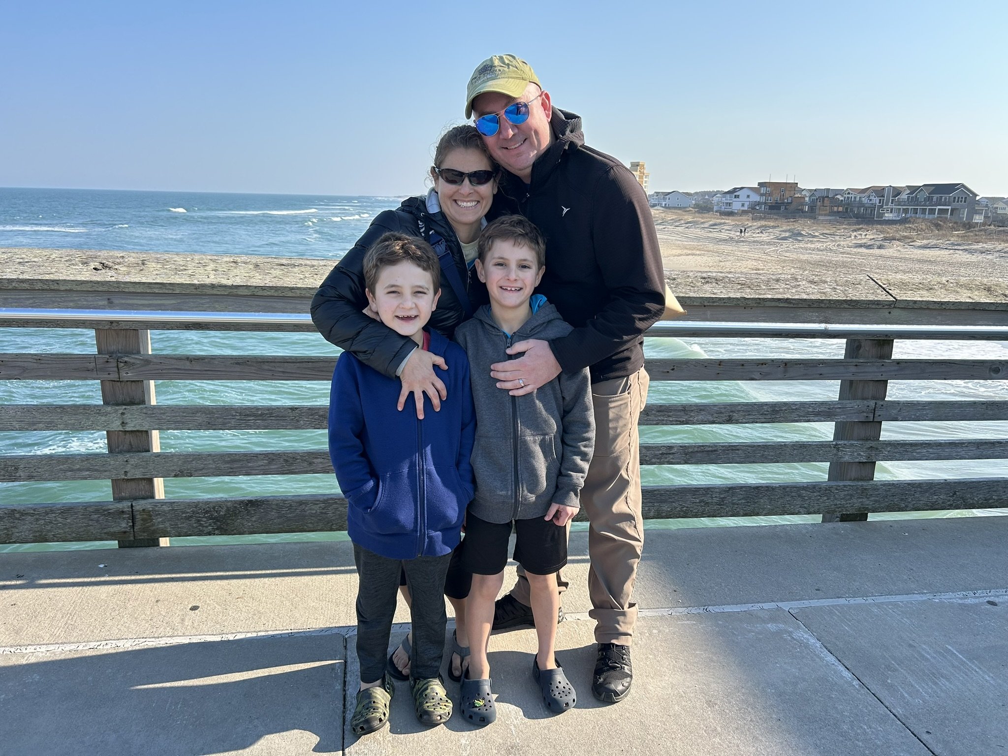 A family of four at the beach, standing on a pier with ocean and houses in the background. The man and woman are smiling, with the woman hugging the two boys, all wearing casual clothes and sunglasses.