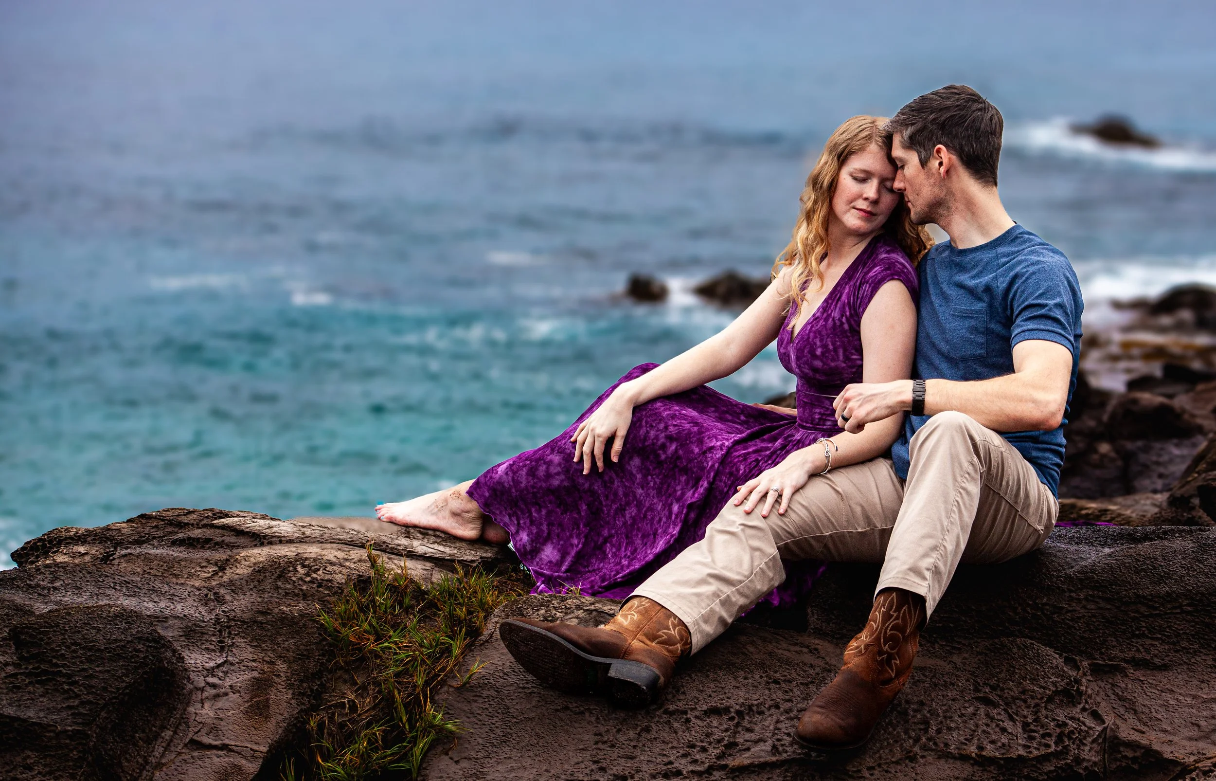 A couple sitting on a rocky shore by the ocean, with their heads touching and eyes closed, appearing peaceful and affectionate.