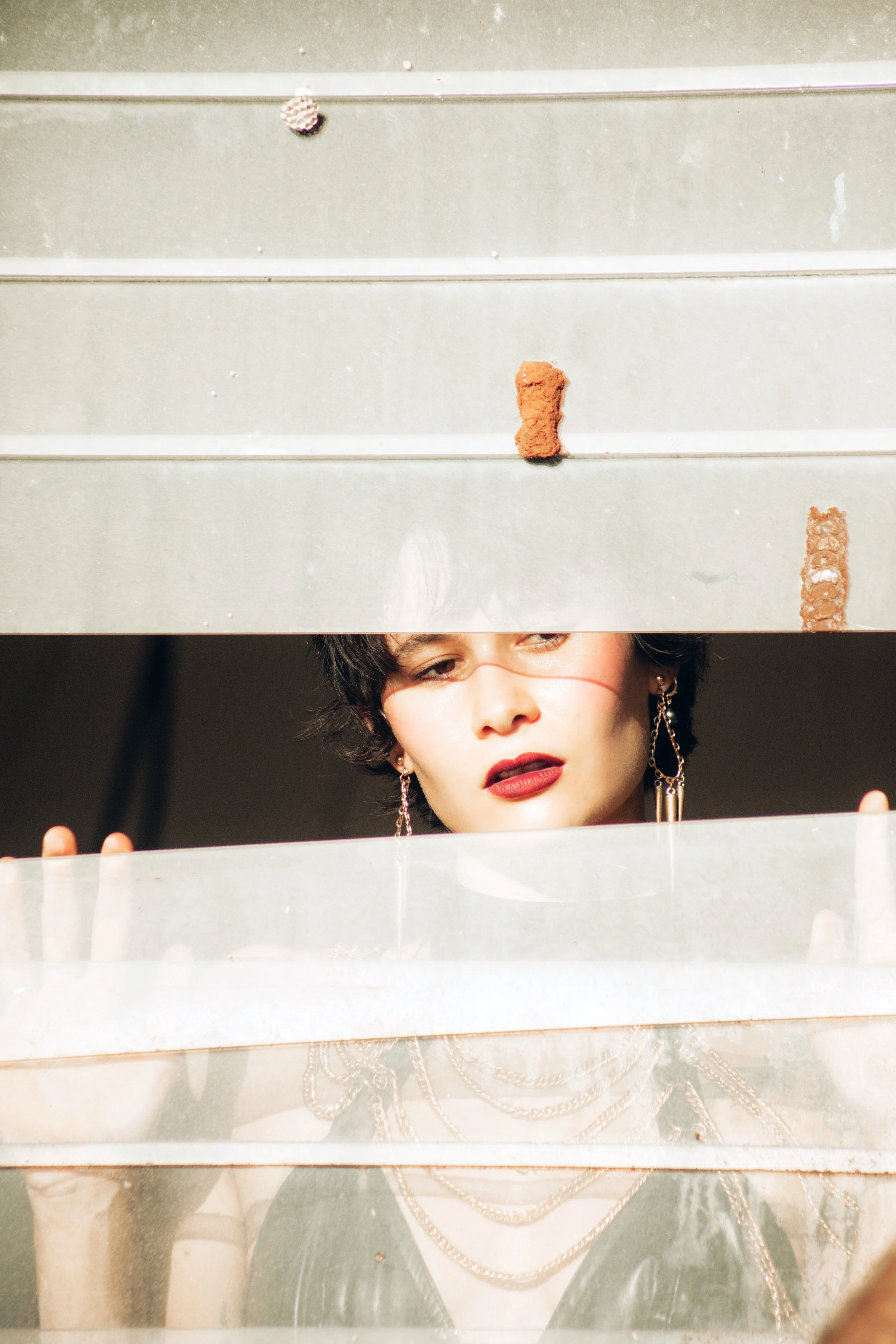 A woman with dark hair, wearing earrings and jewelry, looks through a horizontal gap in a metal surface, with a serious expression on her face.