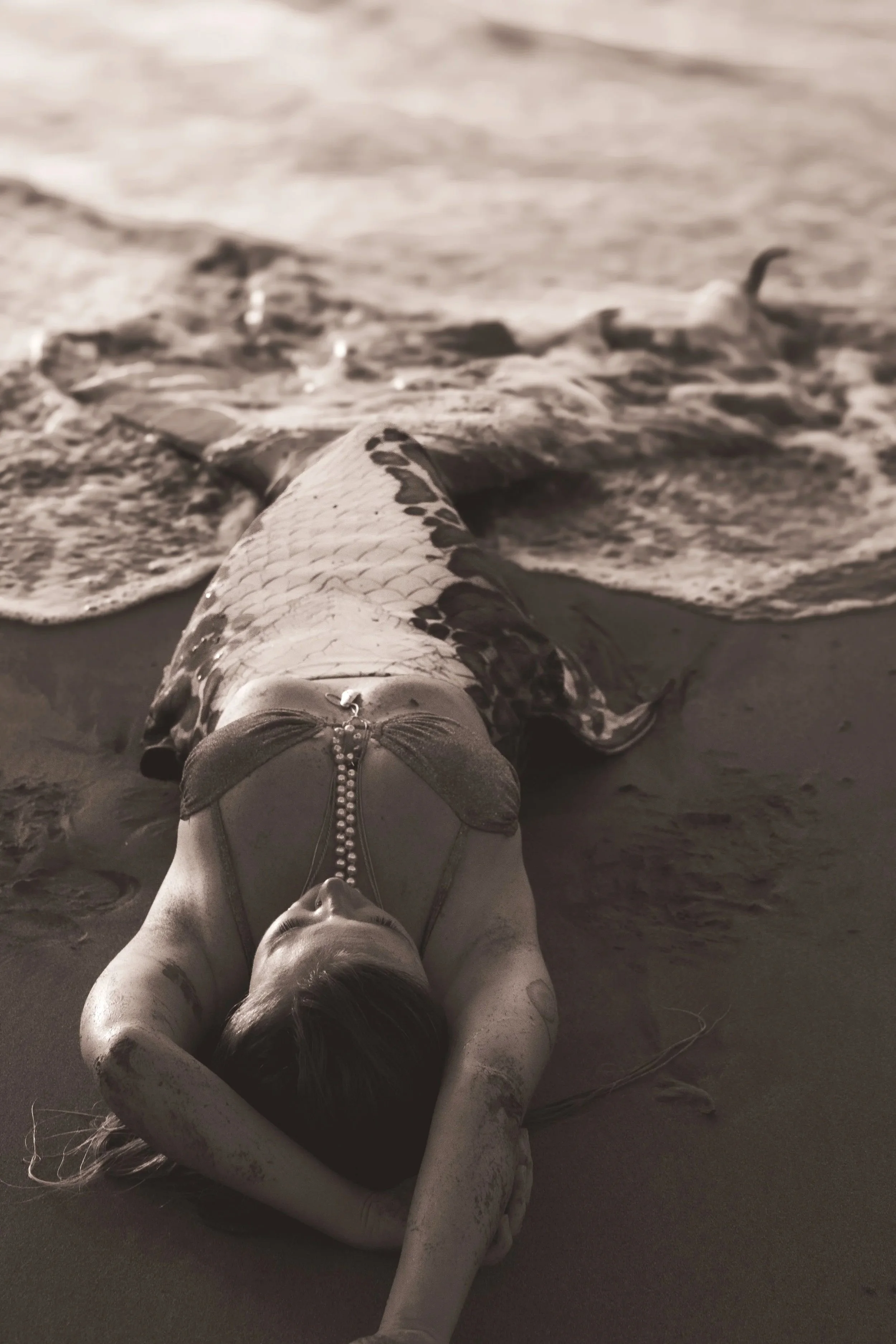 A woman in a swimsuit lying on a sandy beach with her arms above her head, partially covered in sand, as waves wash over her.
