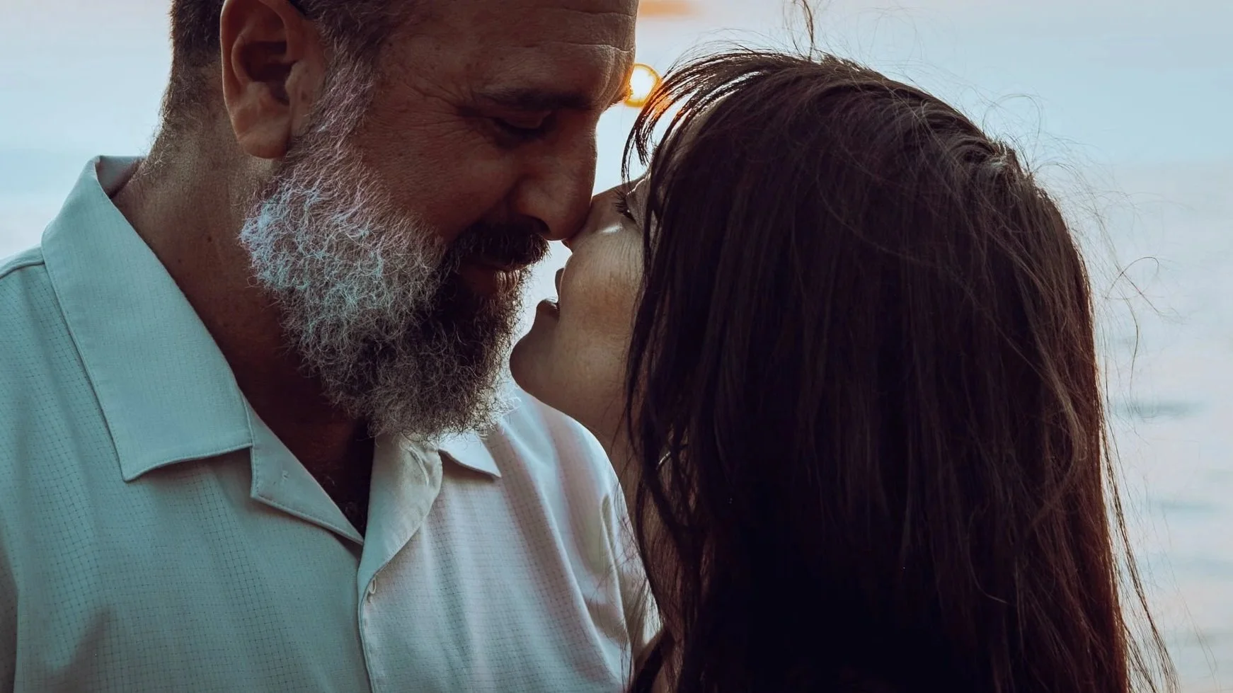 A man and woman are about to kiss, with the man on the left and the woman on the right, at the beach during sunset.
