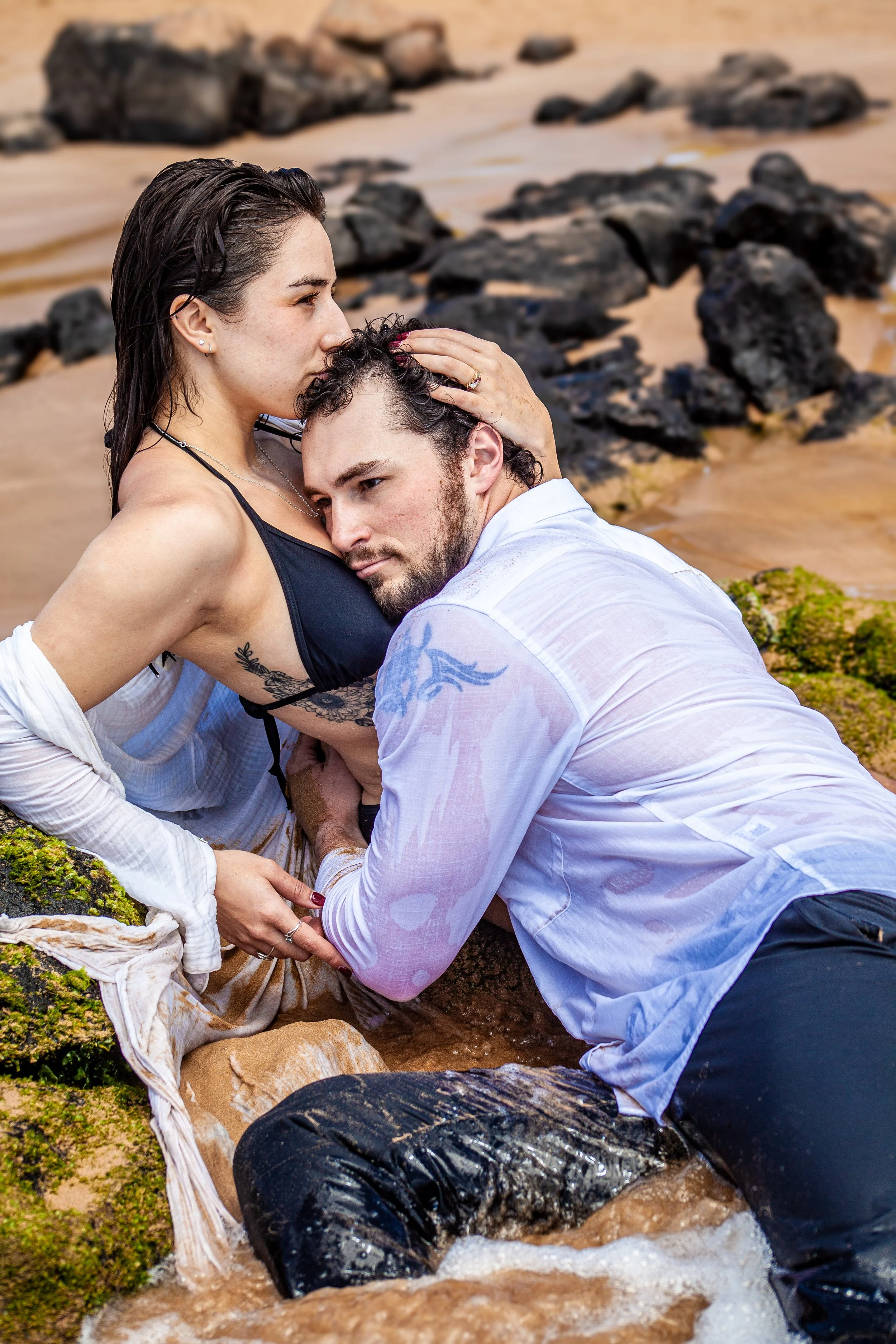 A woman in a black bikini top and a man in a white shirt are embracing on a rocky beach, with water and rocks in the background.
