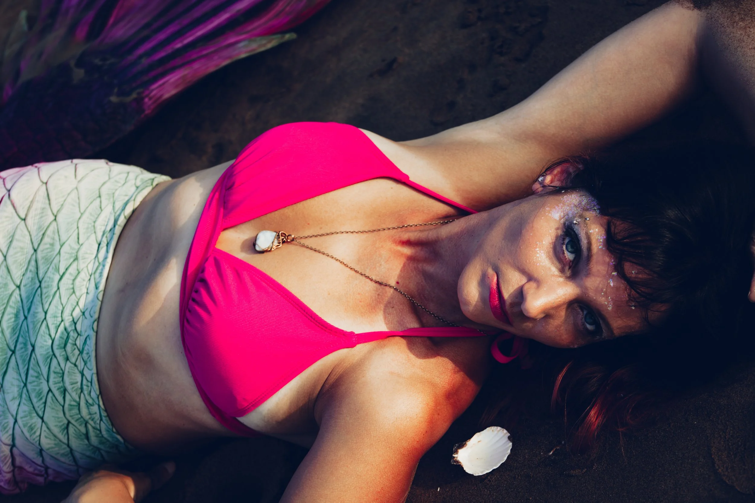 Woman lying on dark sand, wearing a bright pink top and a white skirt with green leaf patterns, with glitter on her face and shell accessories, looking towards the camera.