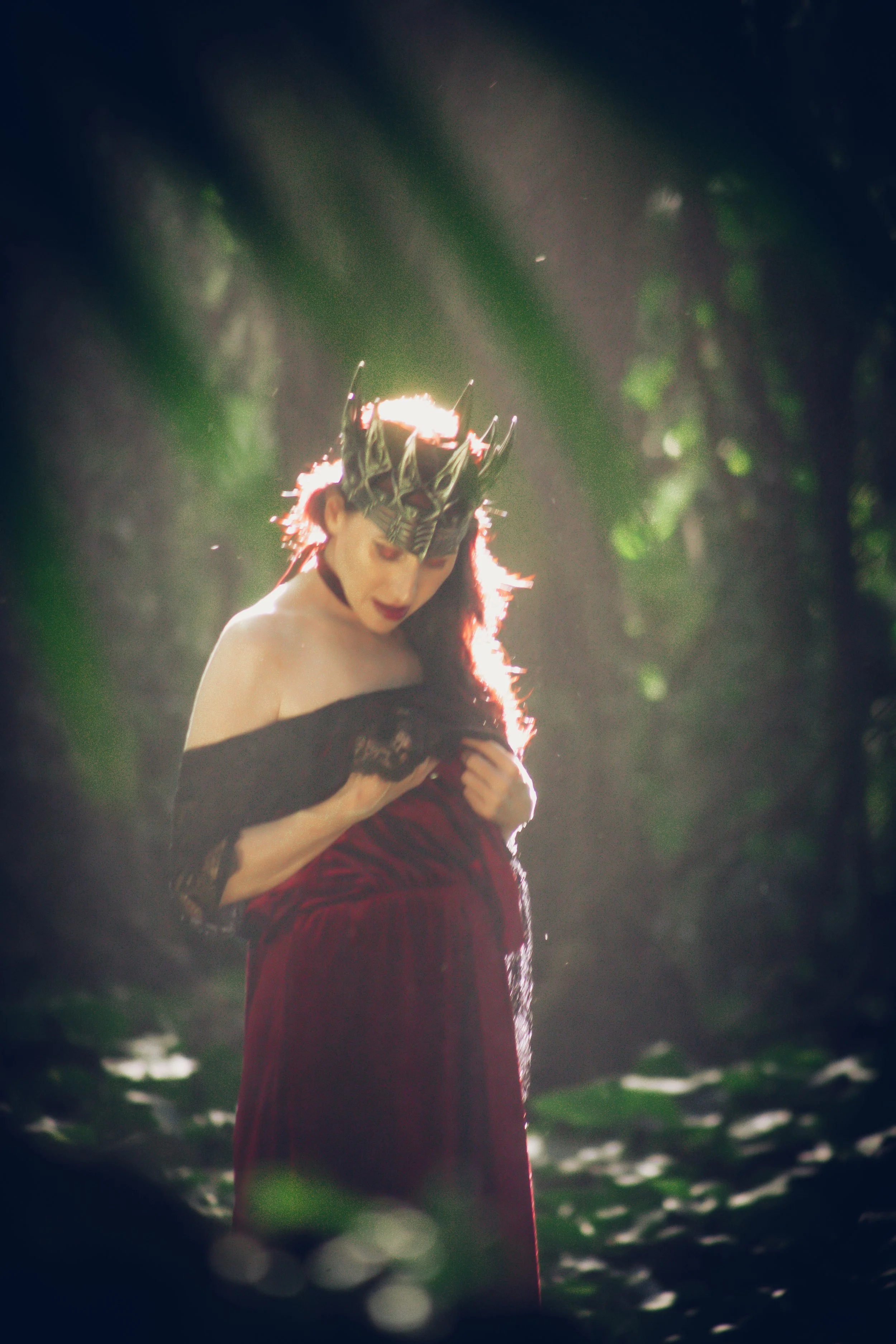 A person dressed as a witch with a black crown and red velvet gown, standing in a forest with sunlight filtering through trees.