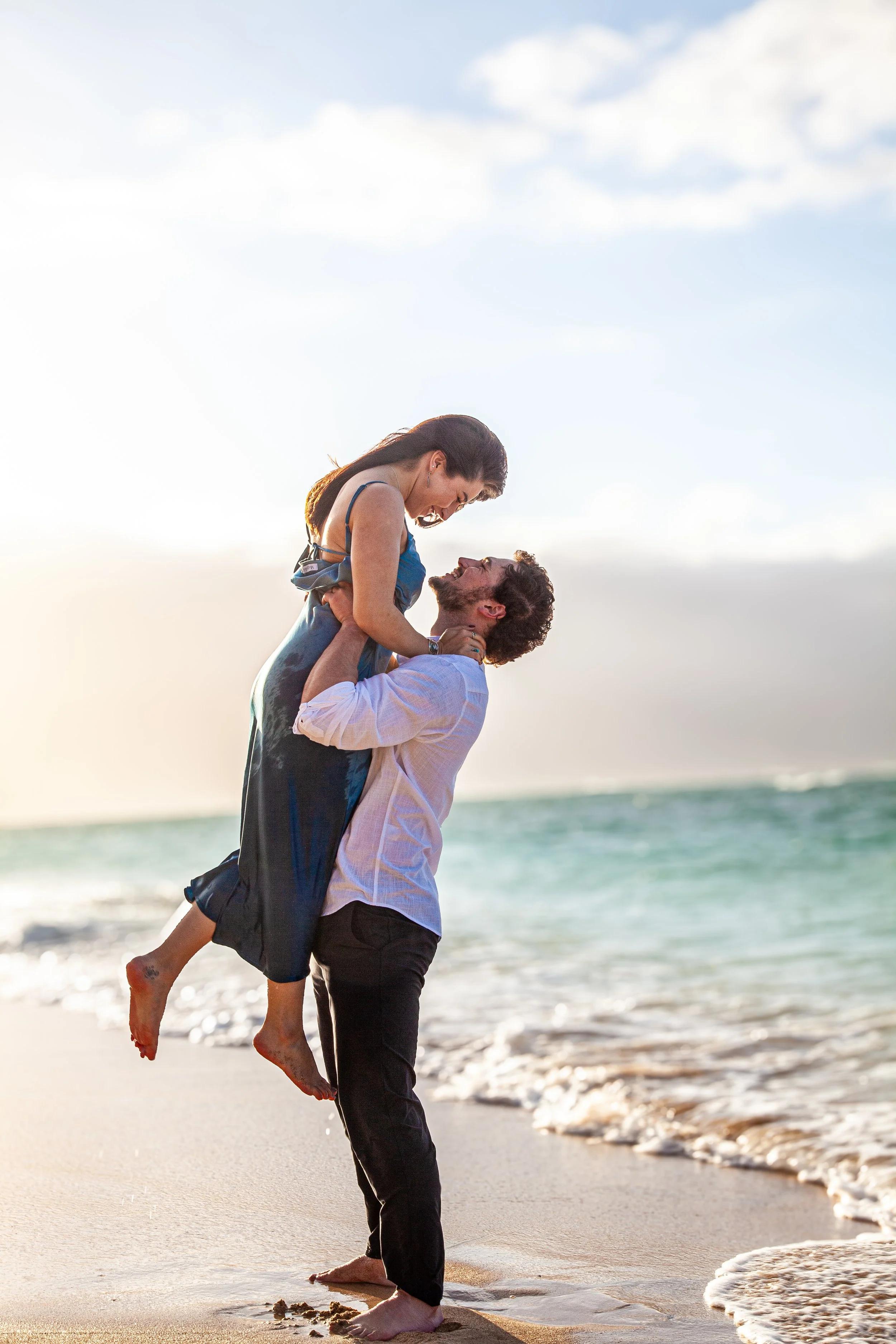 A couple on the beach, the man lifting the woman in his arms, both smiling and looking at each other, with the ocean and sky in the background.