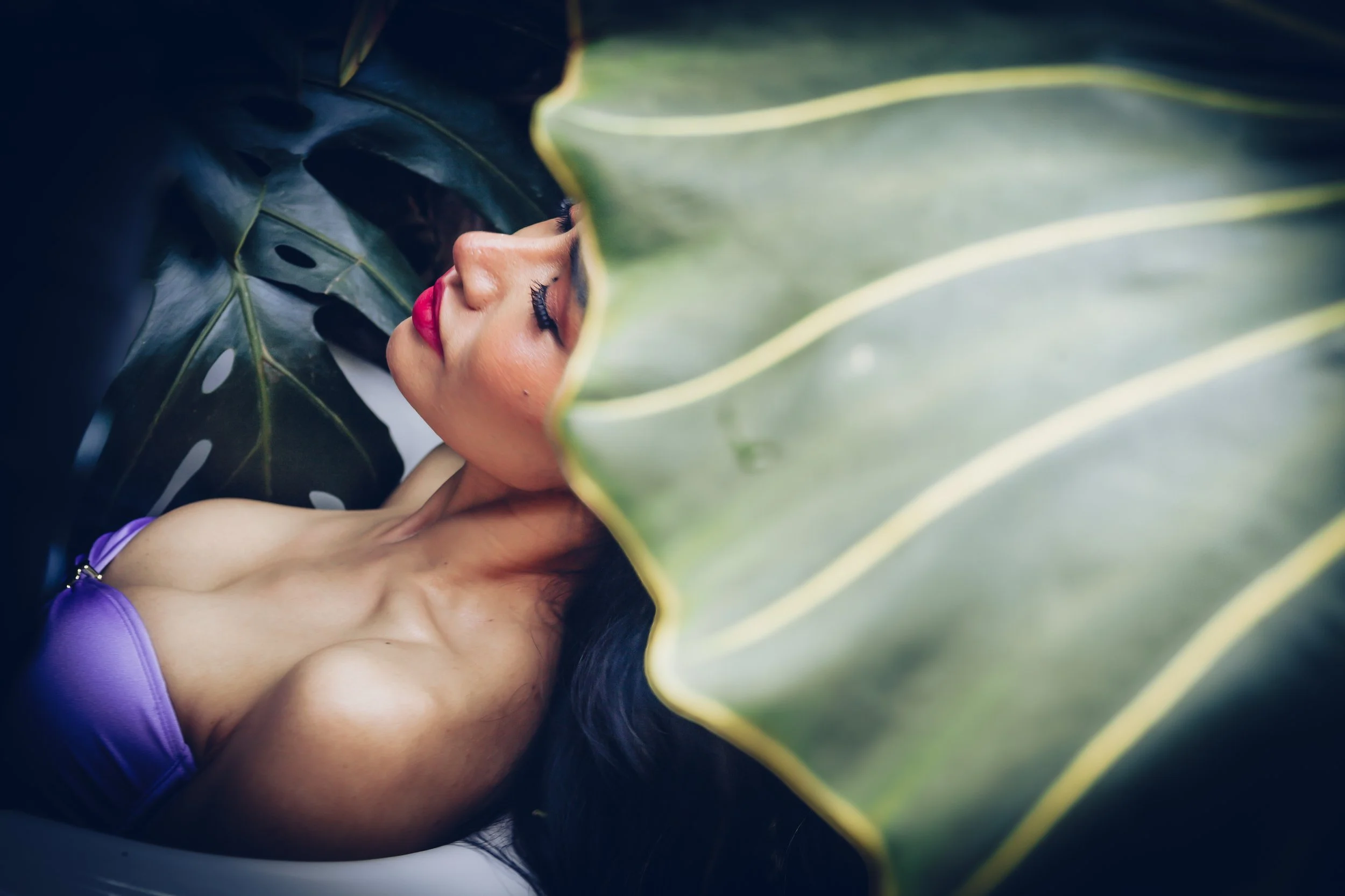 A woman with long black hair and red lipstick sleeping among large green tropical leaves.