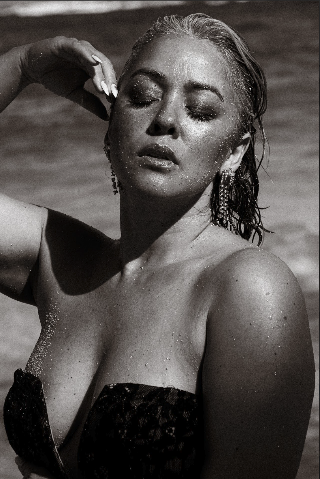 A woman with wet hair and her eyes closed, wearing large earrings, and a strapless lace top, with water droplets on her face and body, standing near water.