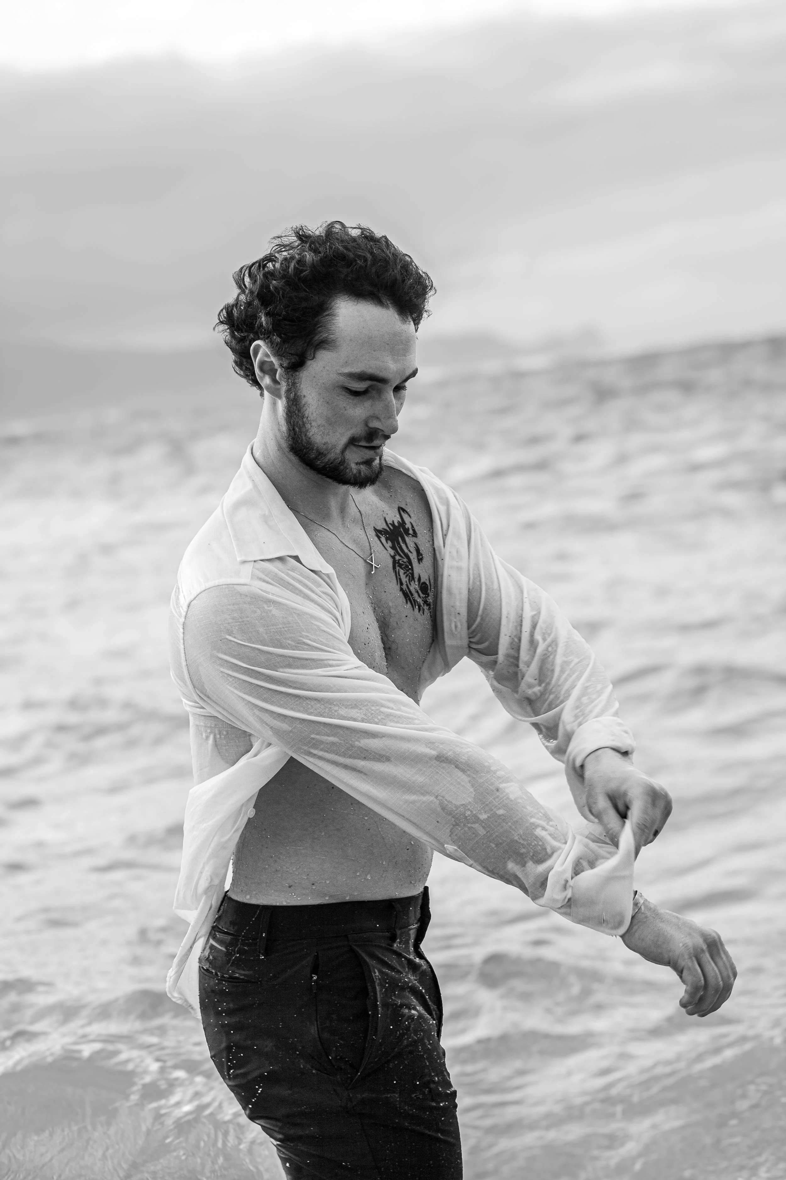 A man with wet hair and a beard in a partially unbuttoned white shirt standing in the ocean, looking down and adjusting his rolled-up sleeve, with a cloudy sky in the background.