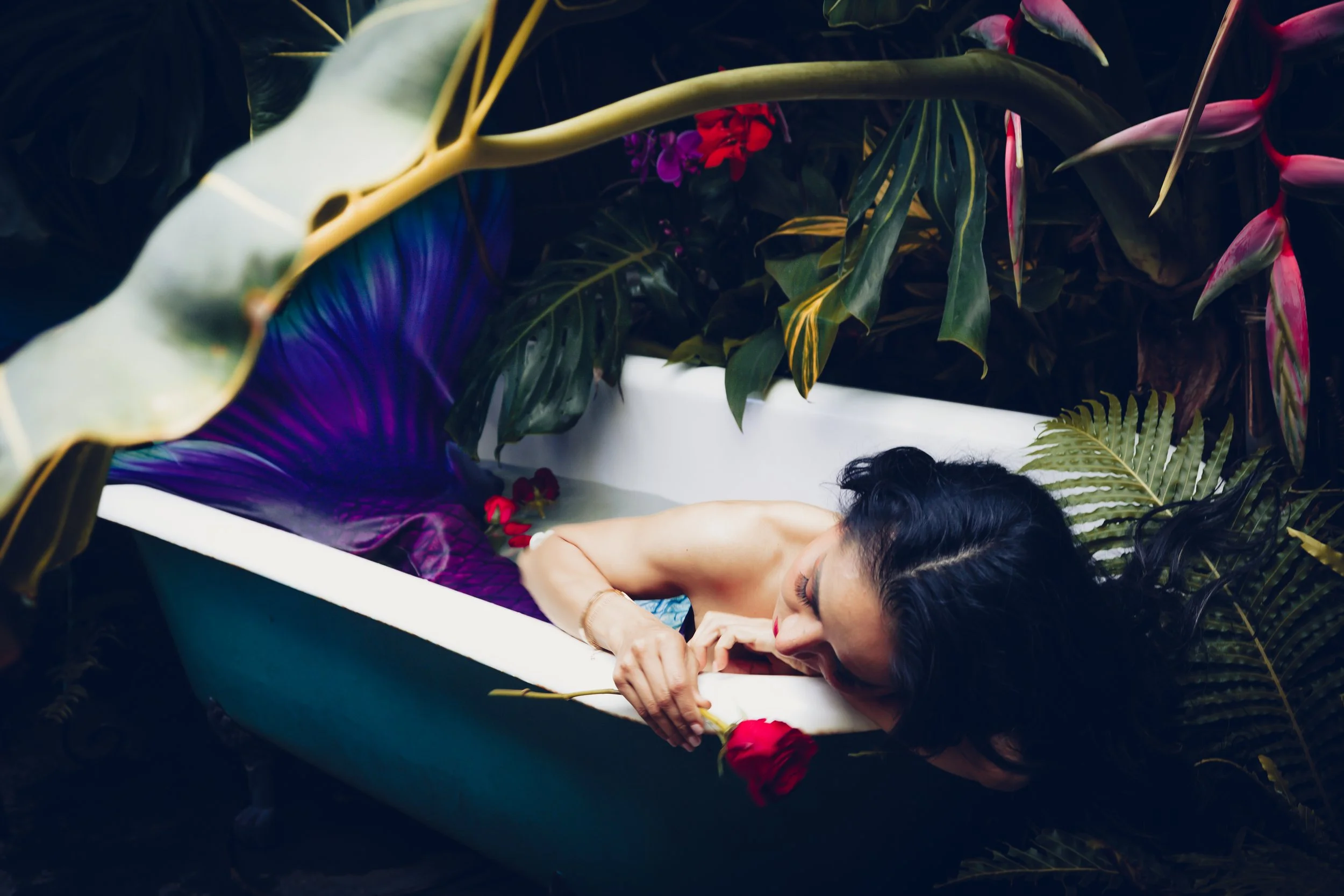 A woman lying in a bathtub surrounded by lush tropical plants, holding a red rose.