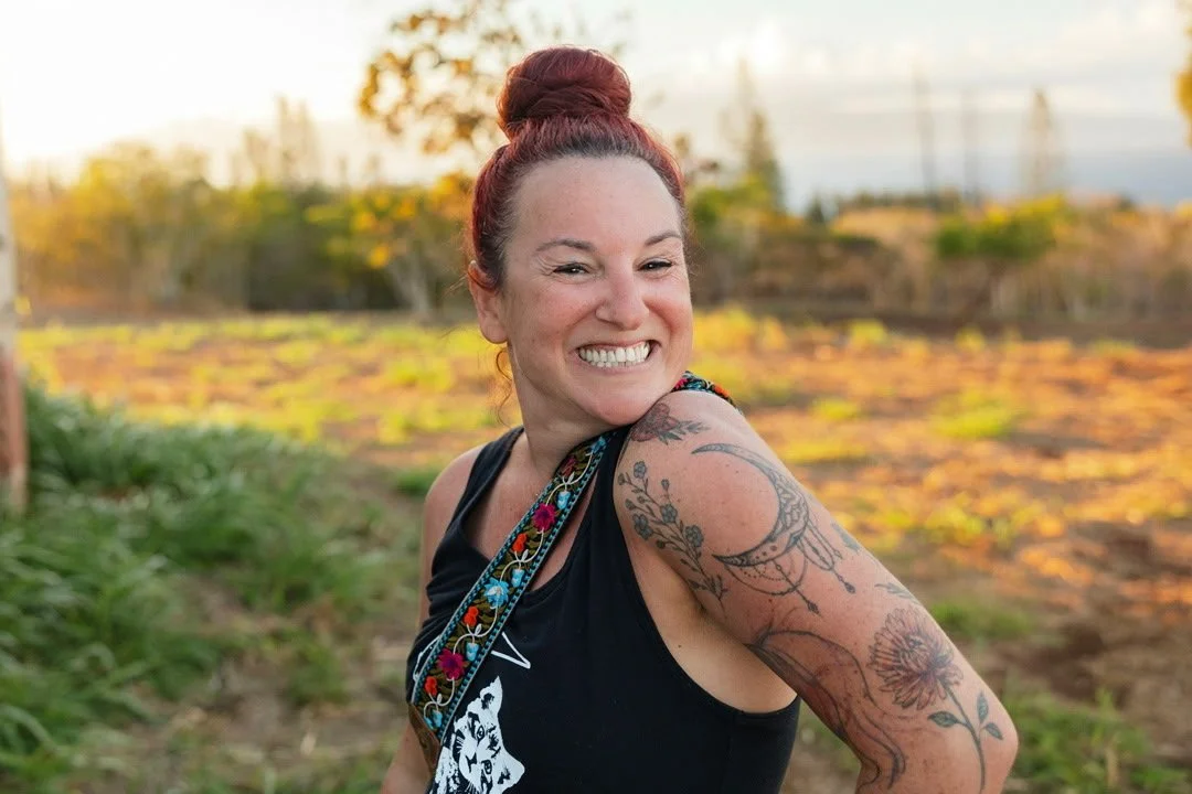 A woman with red hair in a bun smiling outdoors, showing tattoos on her arm, wearing a black tank top and a colorful strap over her shoulder.