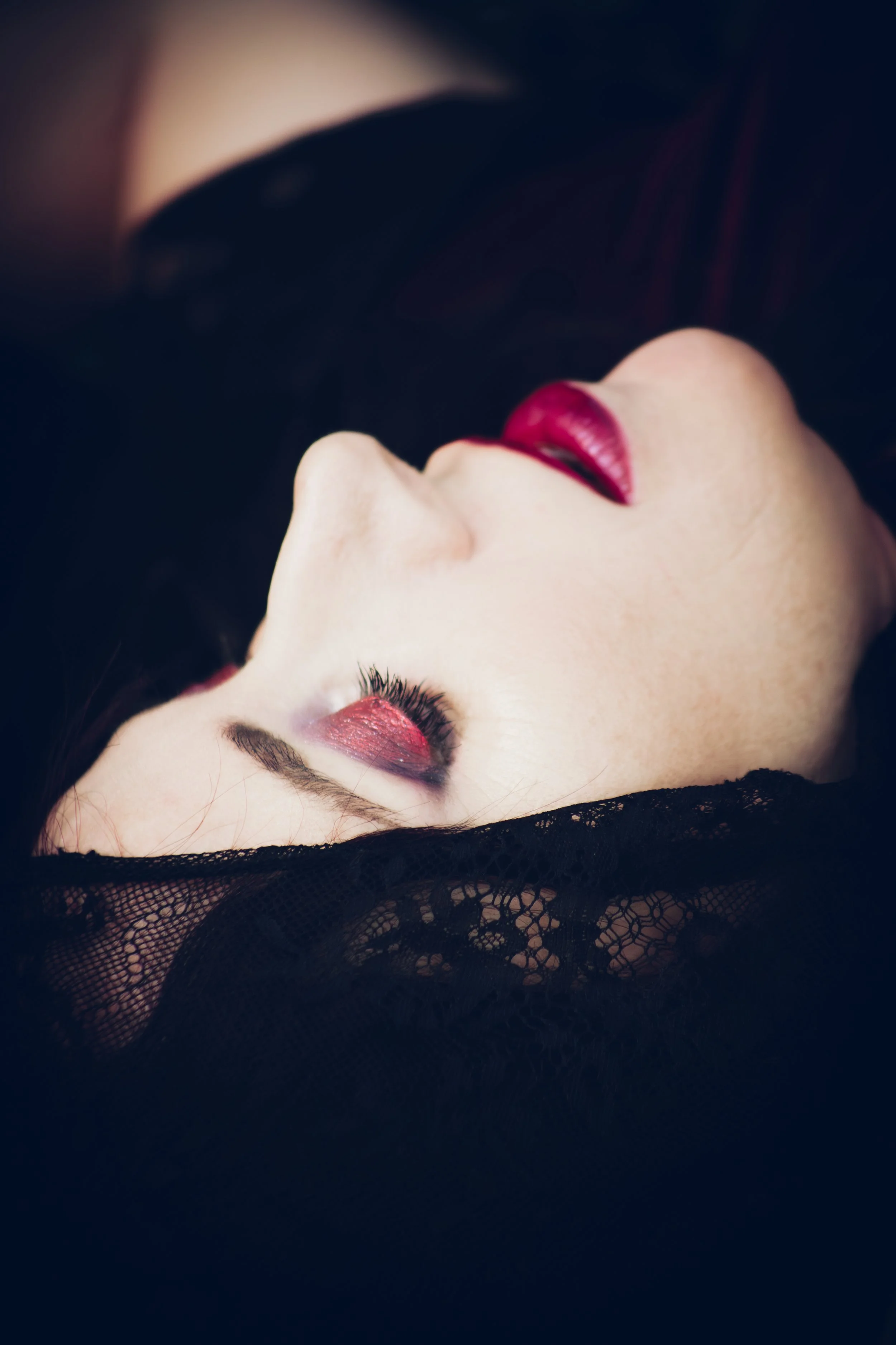 Close-up of a woman's face with red lipstick, lying down with a black lace fabric partially covering her face, soft lighting enhancing her facial features.
