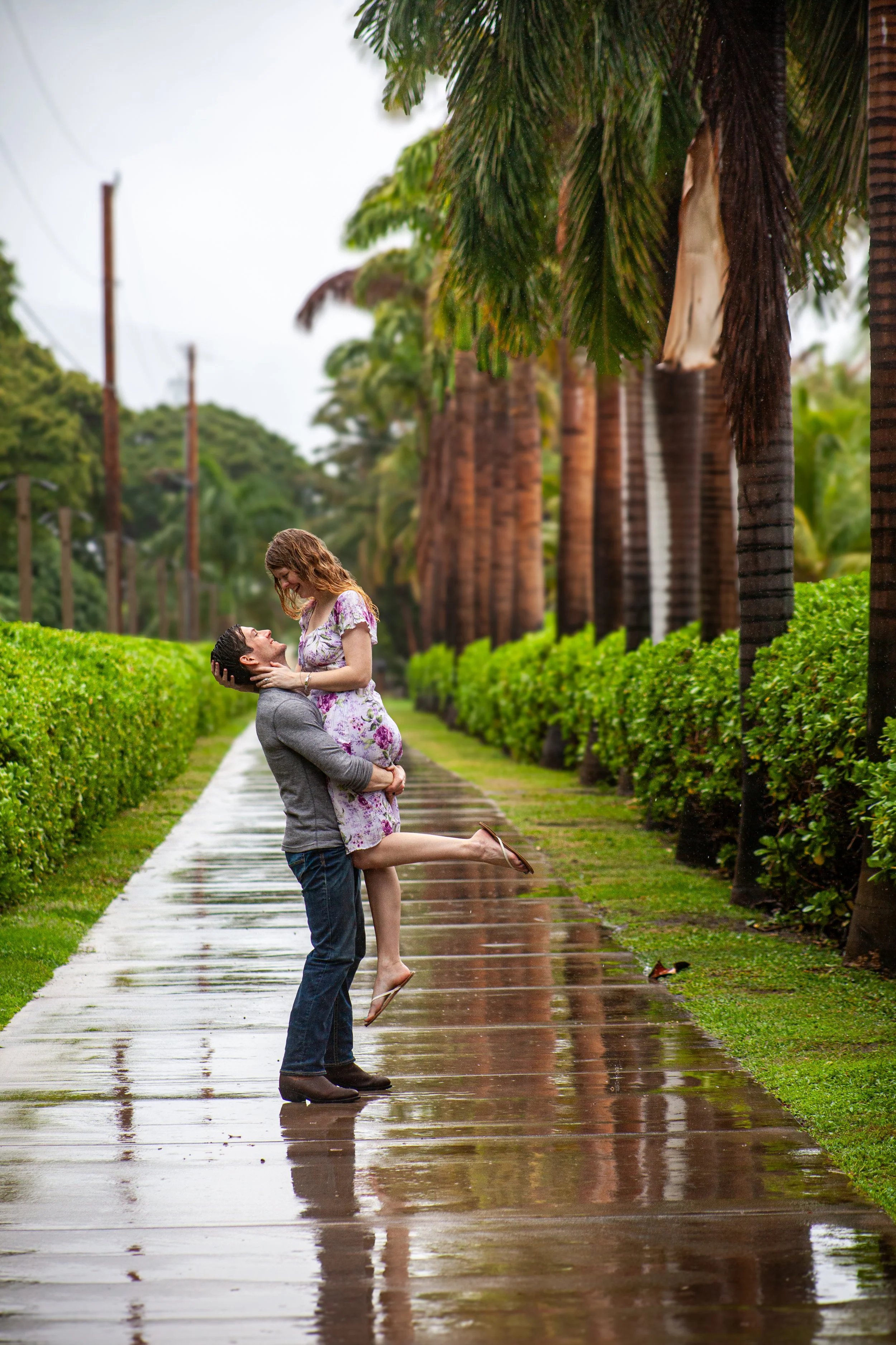 A couple on a rainy day, with the man lifting the woman in his arms, standing on a wet sidewalk lined with green bushes and tall palm trees.