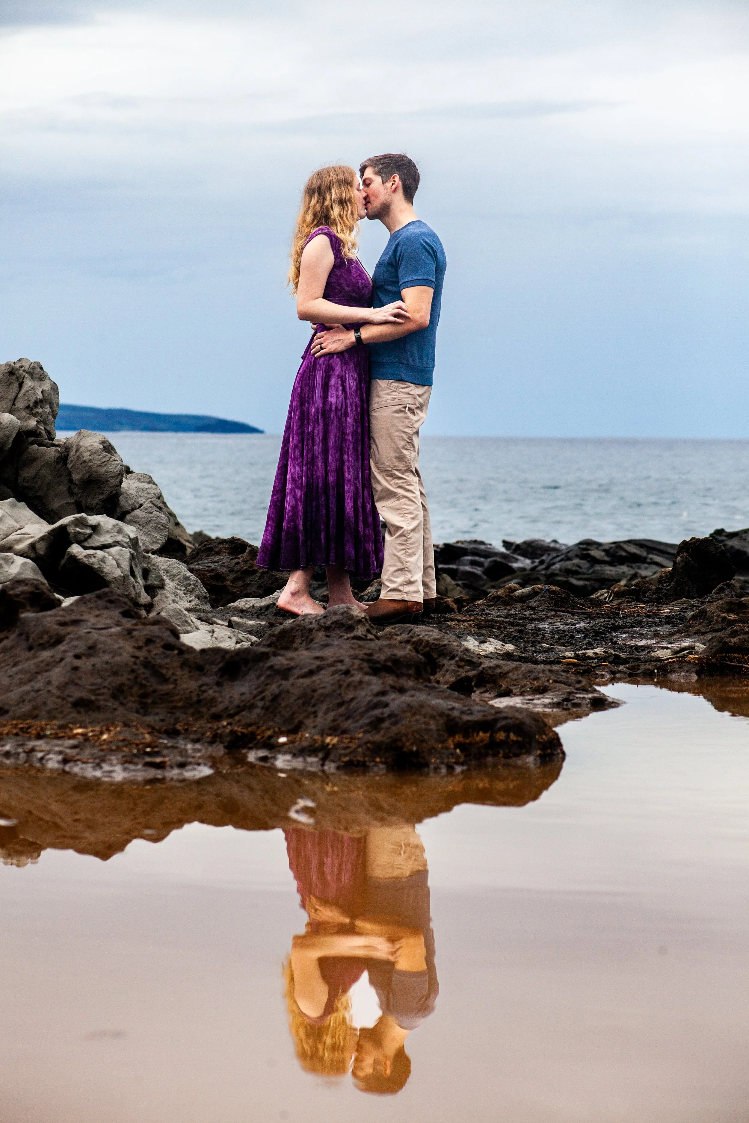 A couple standing on rocks by the water, about to kiss, with their reflection visible in a shallow pool of water.