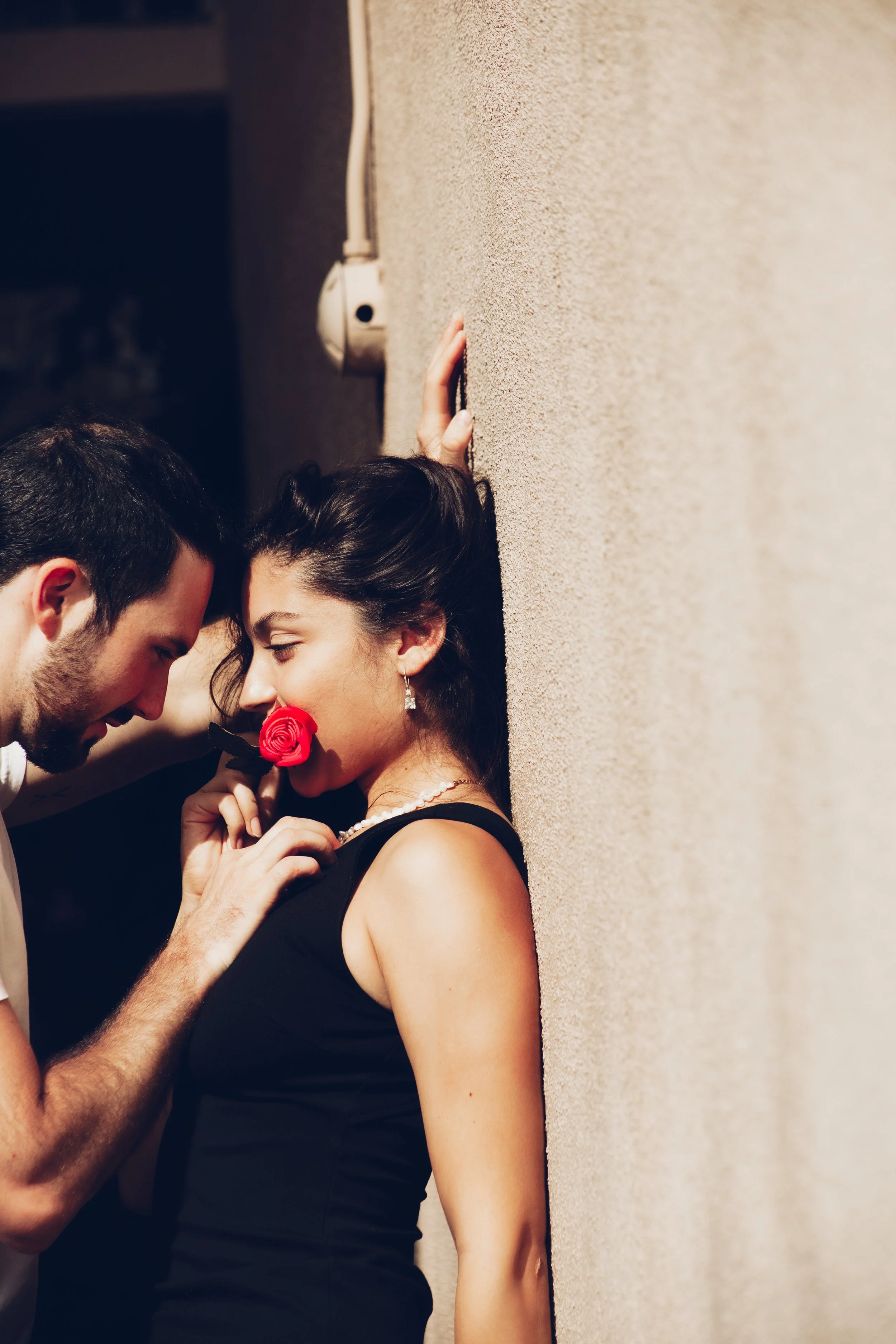 A man and woman are in a romantic scene; the man leans in close to the woman, who is holding a red rose in her mouth, while standing against a textured beige wall.
