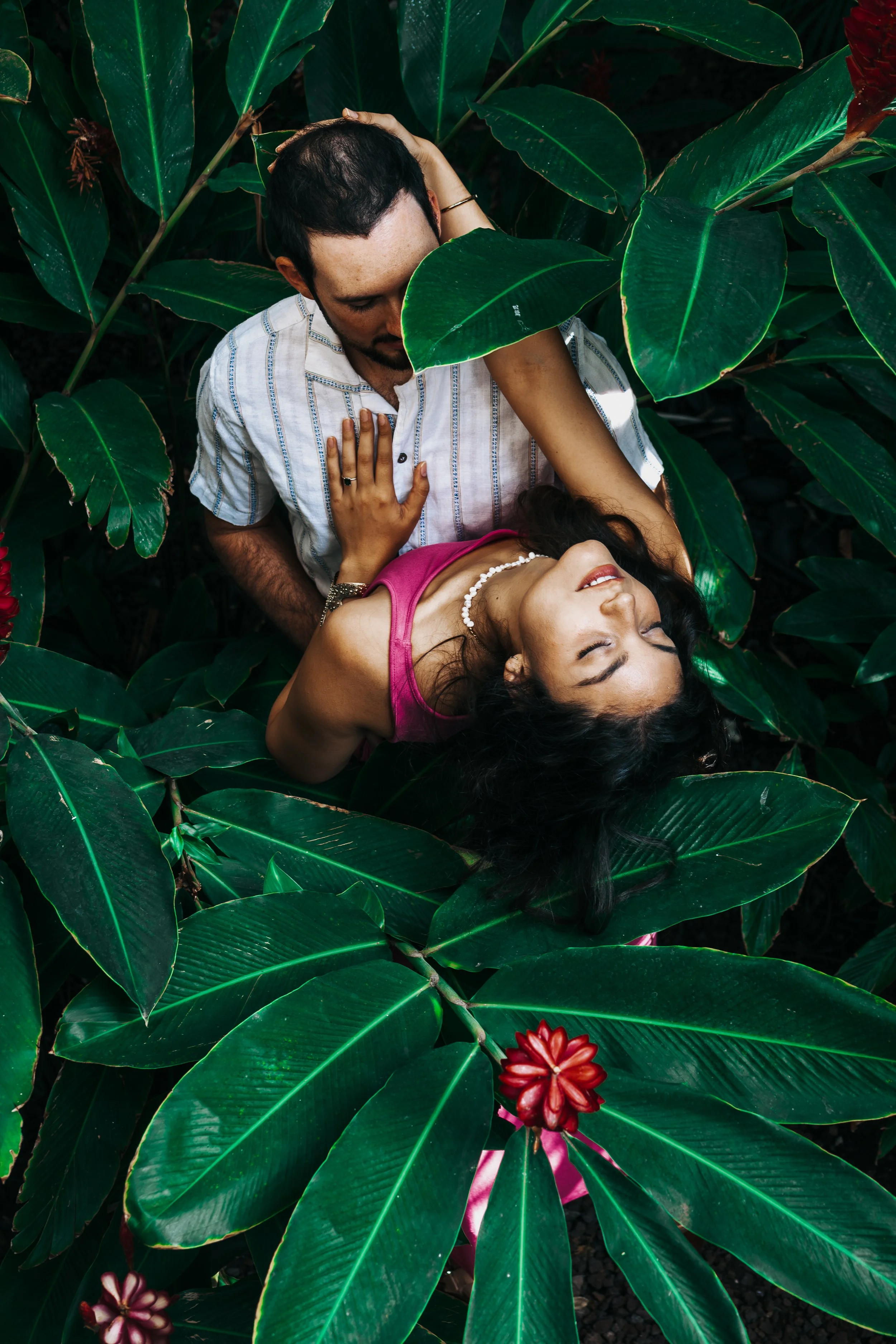 A man and woman are lying among large green leaves, with the woman smiling and eyes closed, wearing a pink dress and pearl necklace, and the man looking down at her with his hand on her chest.