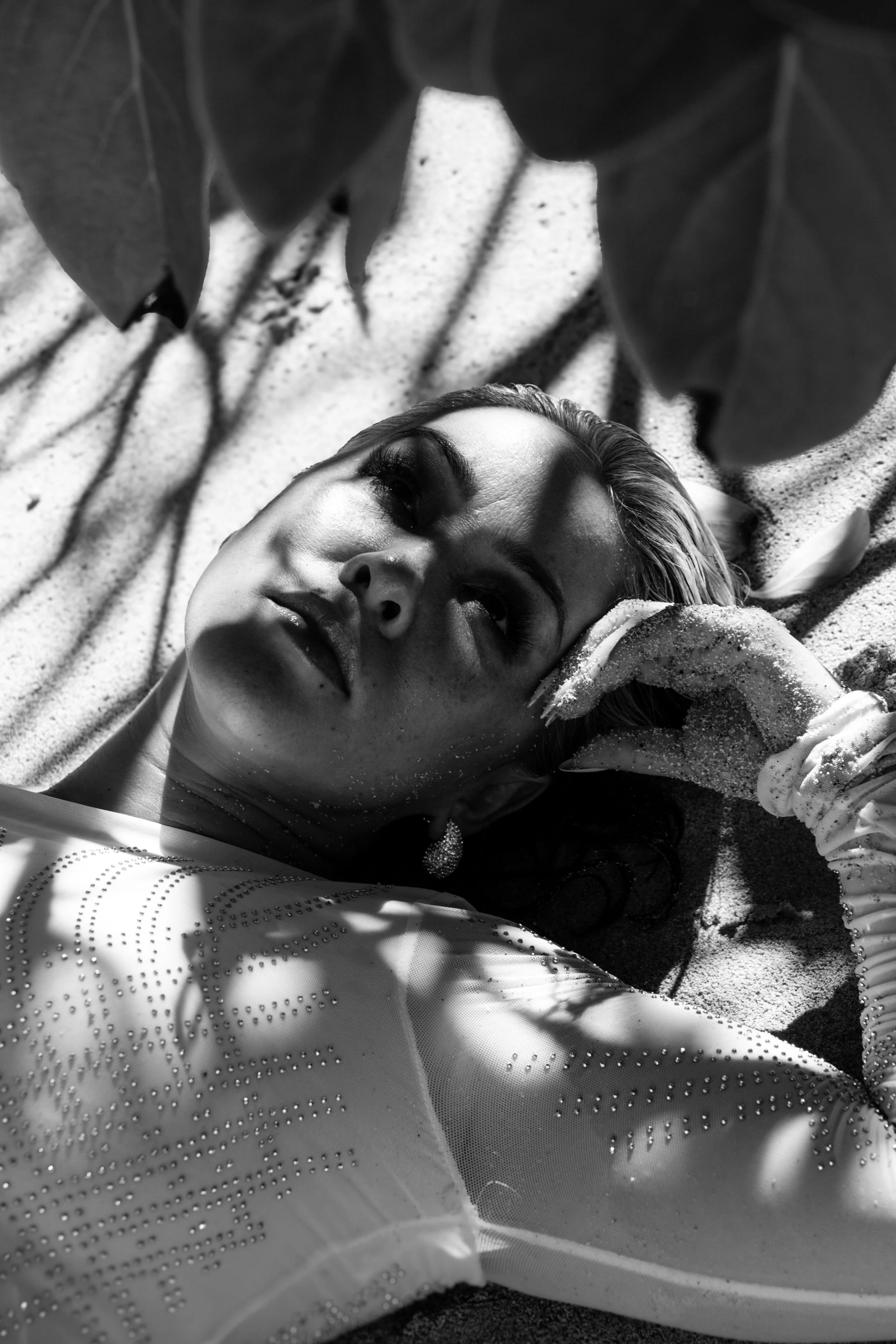 A woman with makeup and jewelry lying on sand with shadows of leaves overhead.