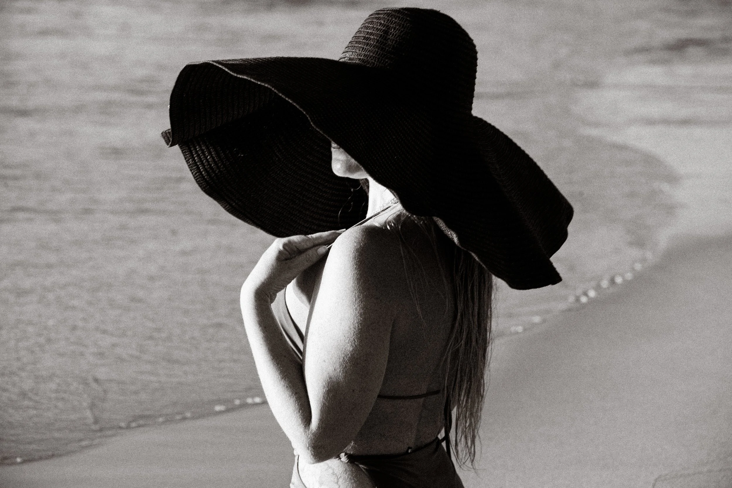 A woman wearing a large sun hat on the beach, standing in shallow water, with her hand resting on her shoulder, in black and white photography.