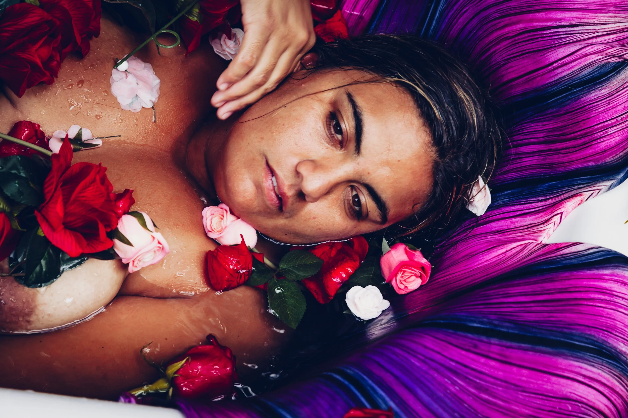 A woman lying in a bath surrounded by and covered with various red, pink, and white roses, with colorful purple, pink, and blue hair.