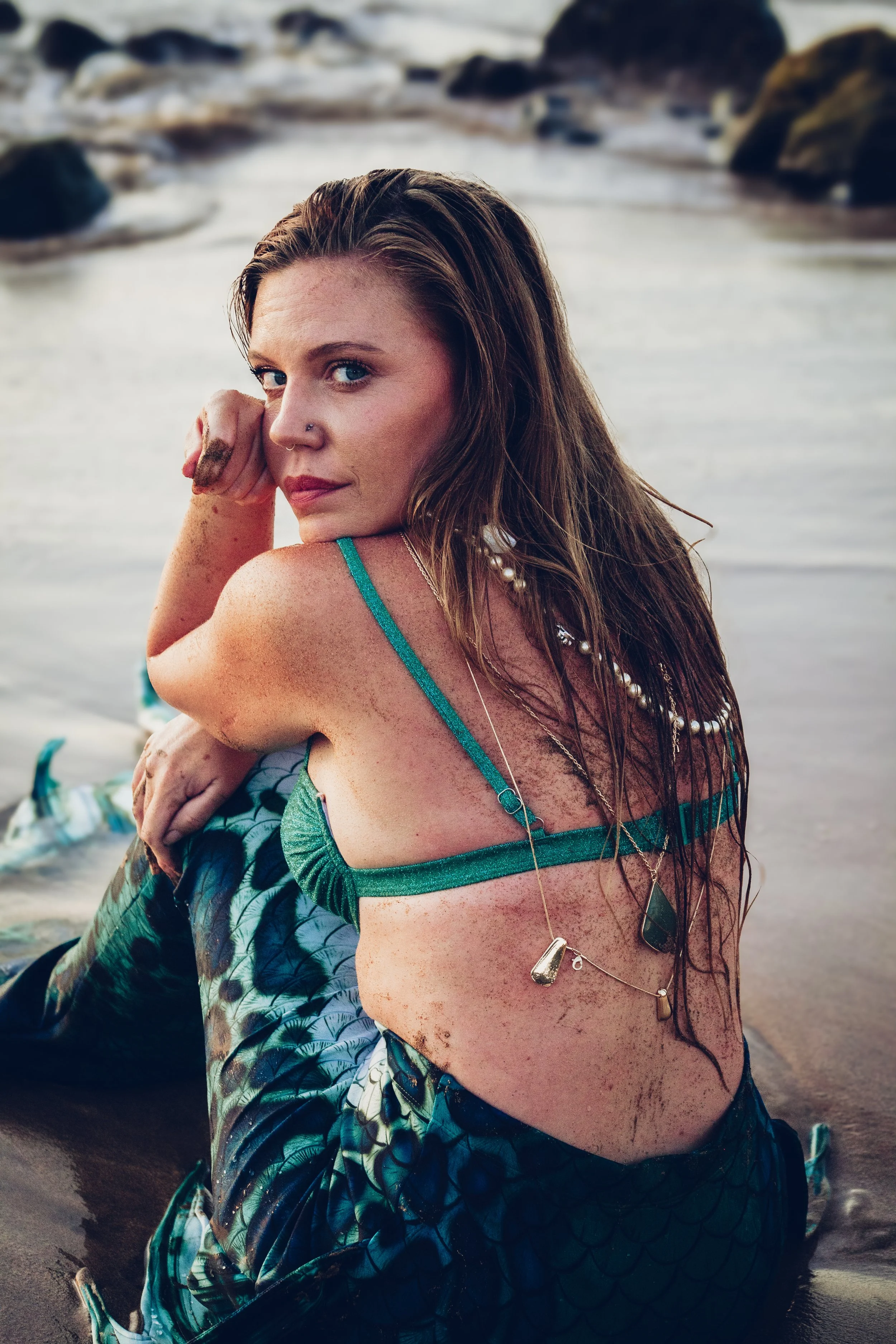 A woman with long hair and blue eyes sitting on the beach in the shallow water, wearing a mermaid costume with a scale pattern, with rocks and water in the background.