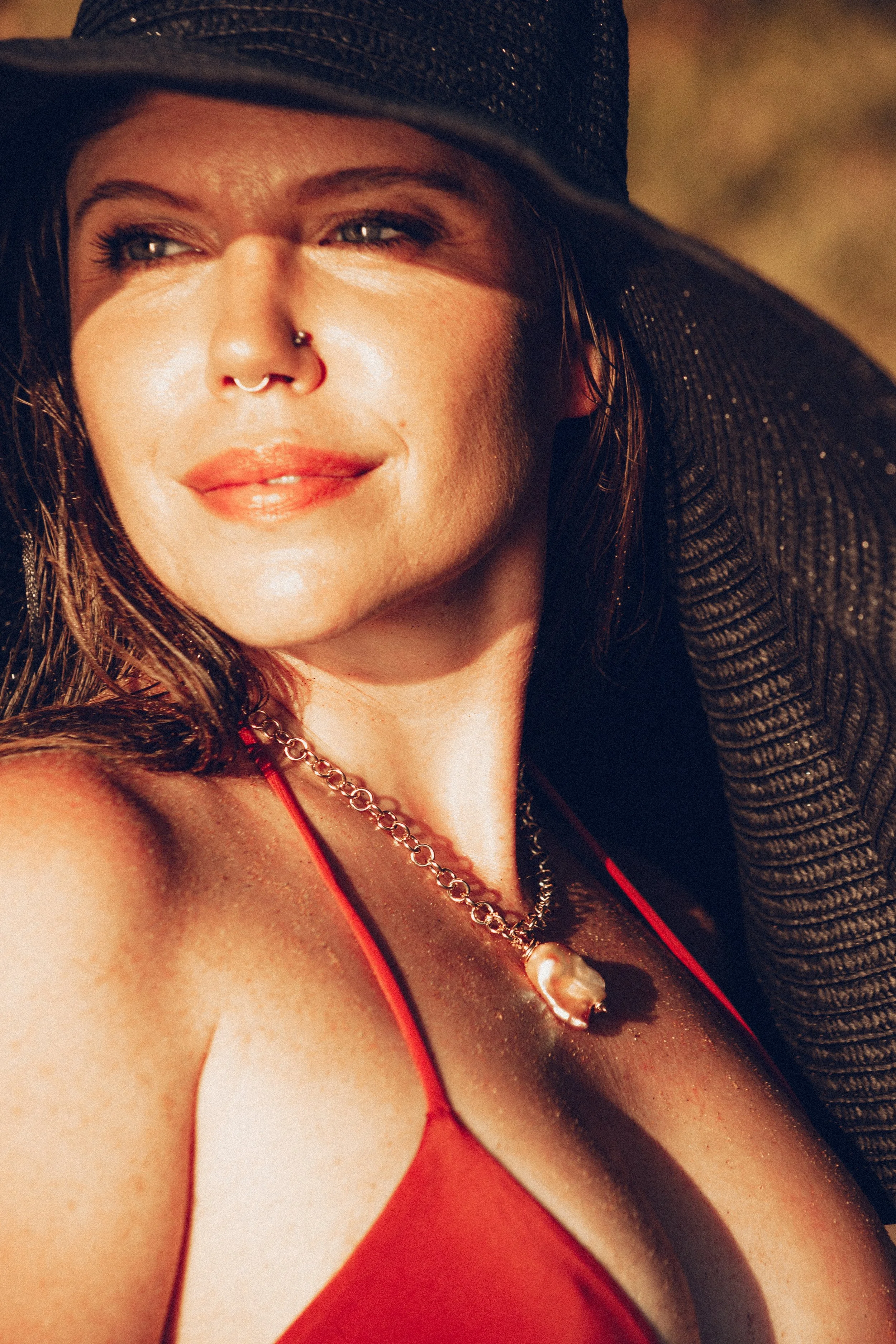 A woman with fair skin wearing a black hat, a red top, and a pearl necklace, sunlight illuminating her face and shoulder.