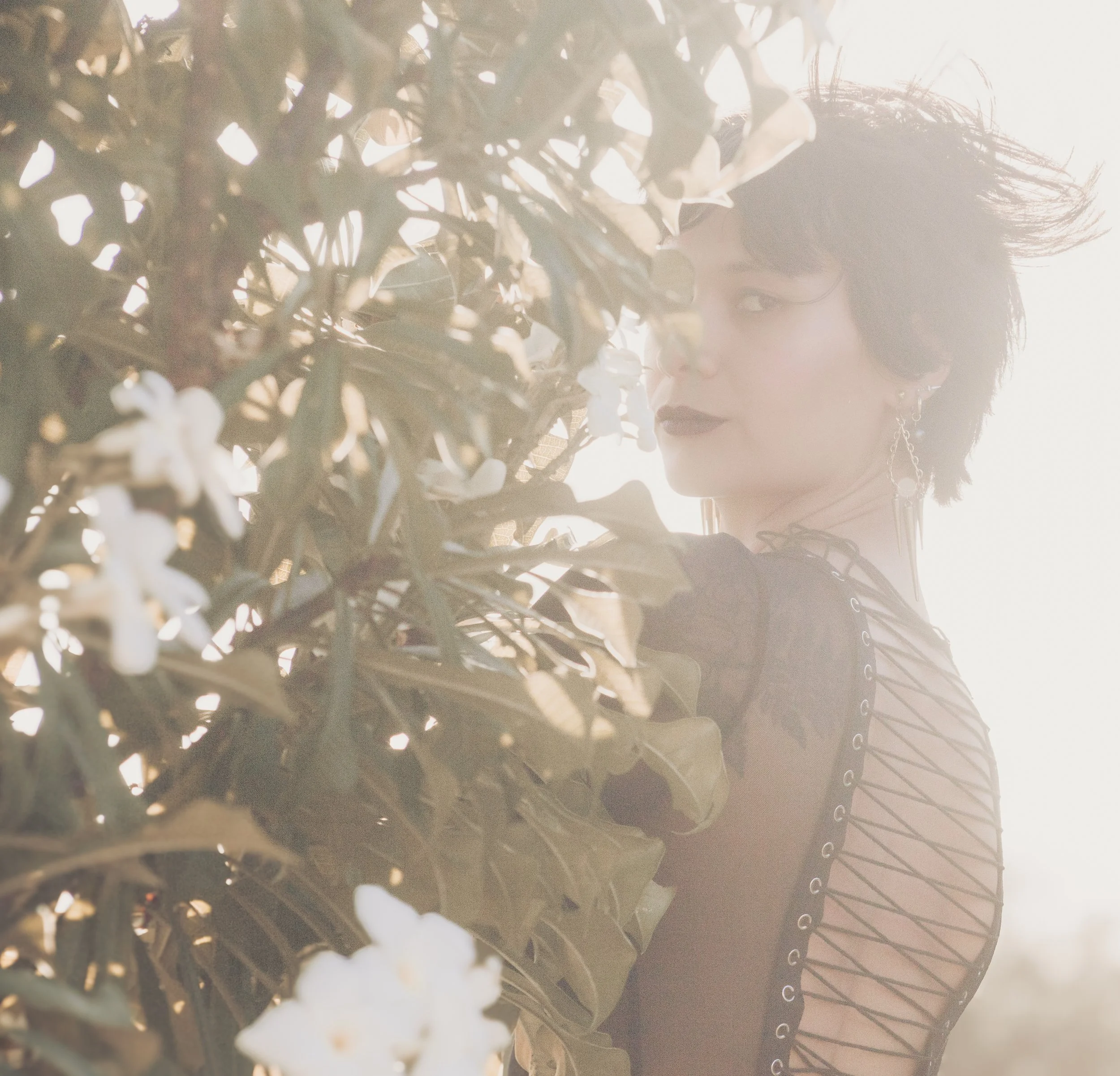 A woman with short dark hair, dark lipstick, and earrings, is partially obscured behind a bush with white flowers, with sunlight creating a backlit effect.