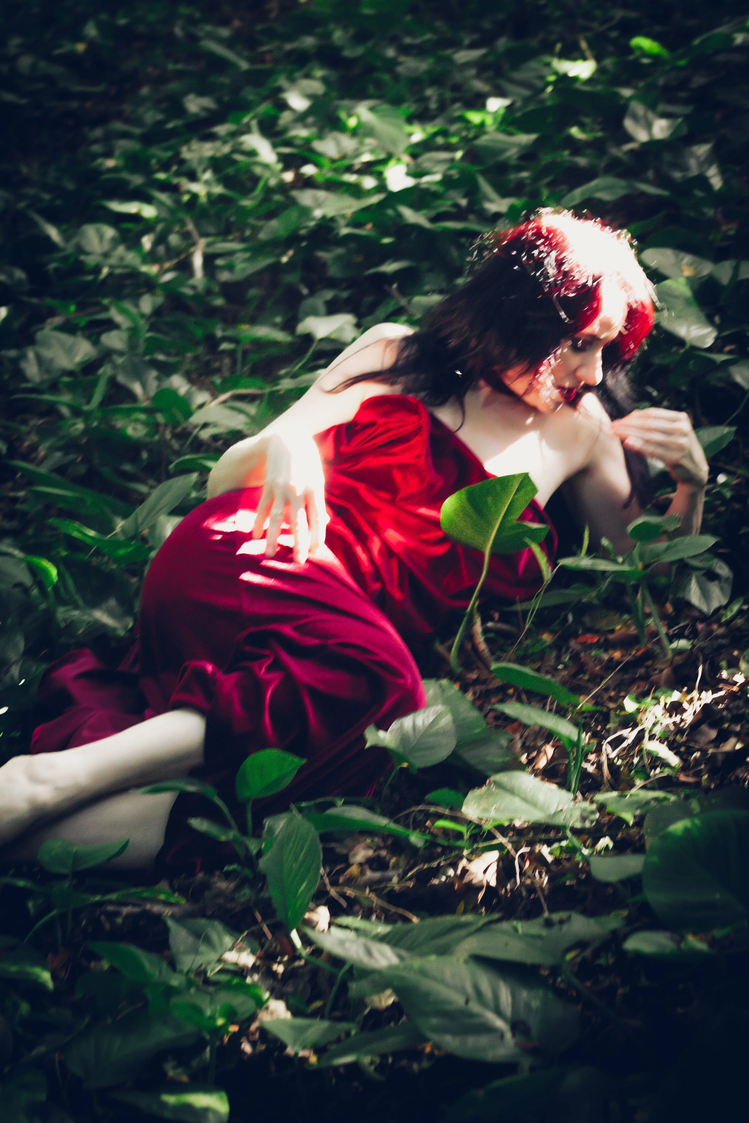 A woman in a red dress laying on the forest floor among green leaves and plants, with sunlight filtering through the foliage.