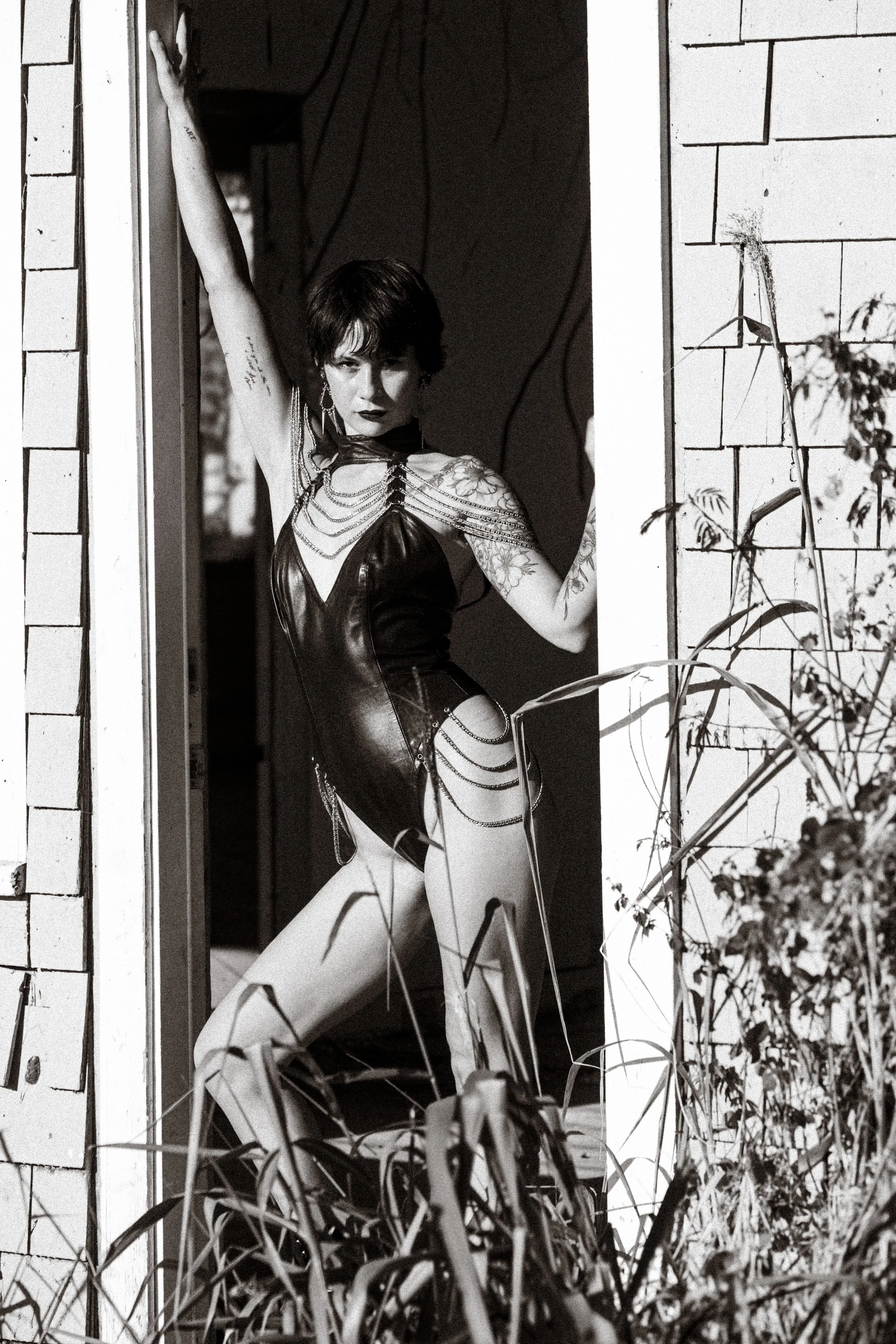 A woman with short dark hair and tattoos on her arms standing in a doorway, wearing a revealing leather dress with chains and jewelry, looking directly at the camera.