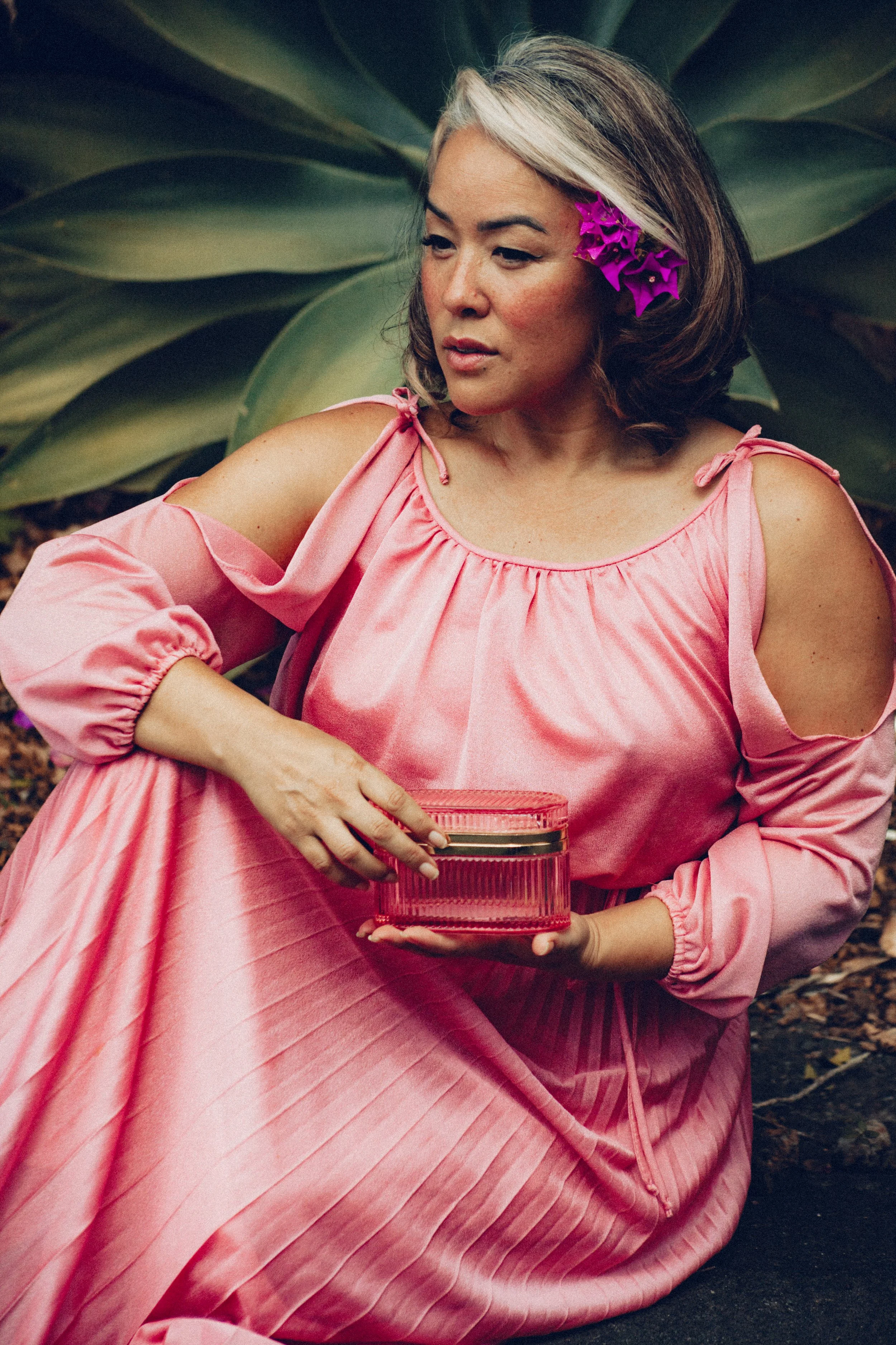 A woman with gray and brown hair wearing a pink dress with cut-out shoulders, holding a pink container, sitting outdoors with large green plants in the background, and a purple flower in her hair.