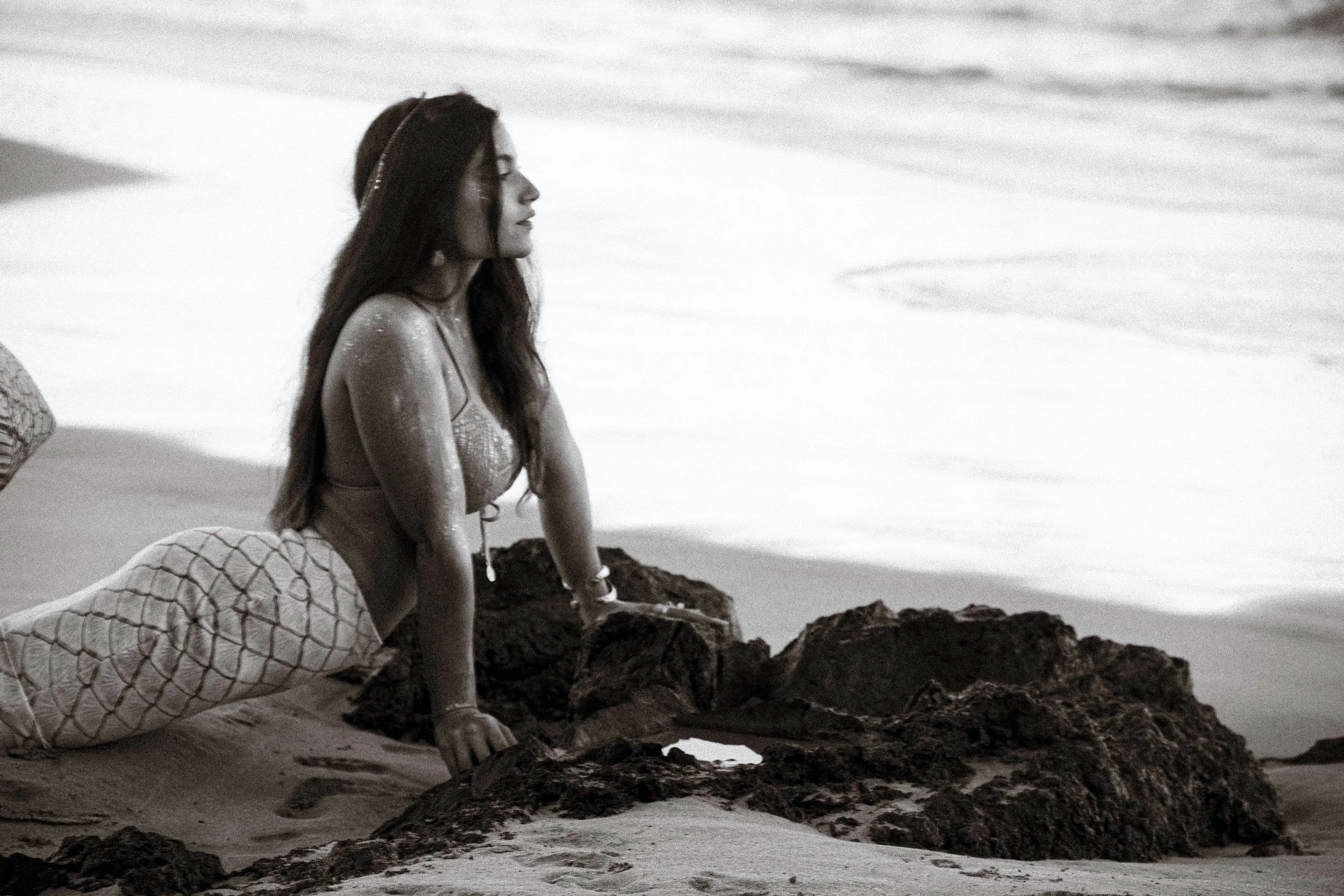 Woman in a swimsuit and fishnet pants kneeling on the beach, leaning on rocks, looking out at the ocean.