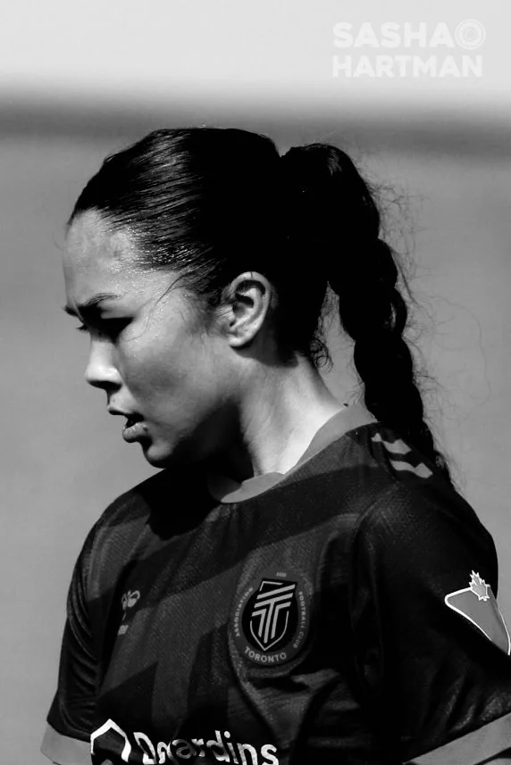 Side profile of a female soccer player with a braid, wearing a AFC Toronto jersey, outdoors.