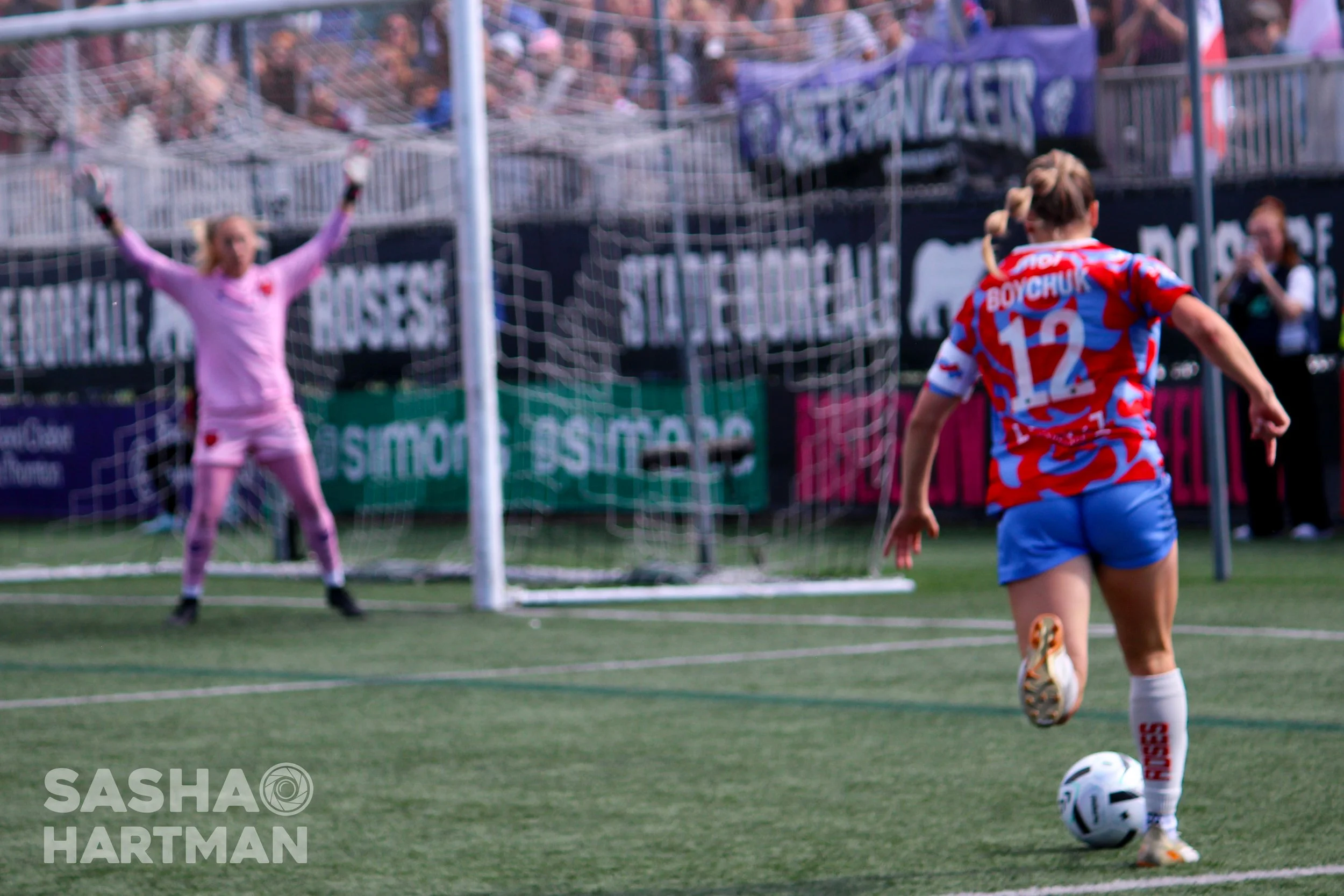 A female soccer player in a red, blue, and white jersey with the number 12 and the name Boychuk, about to kick a ball towards the goal. A goalkeeper in a pink uniform is positioned in front of the goal with arms raised. Spectators are visible in the background.