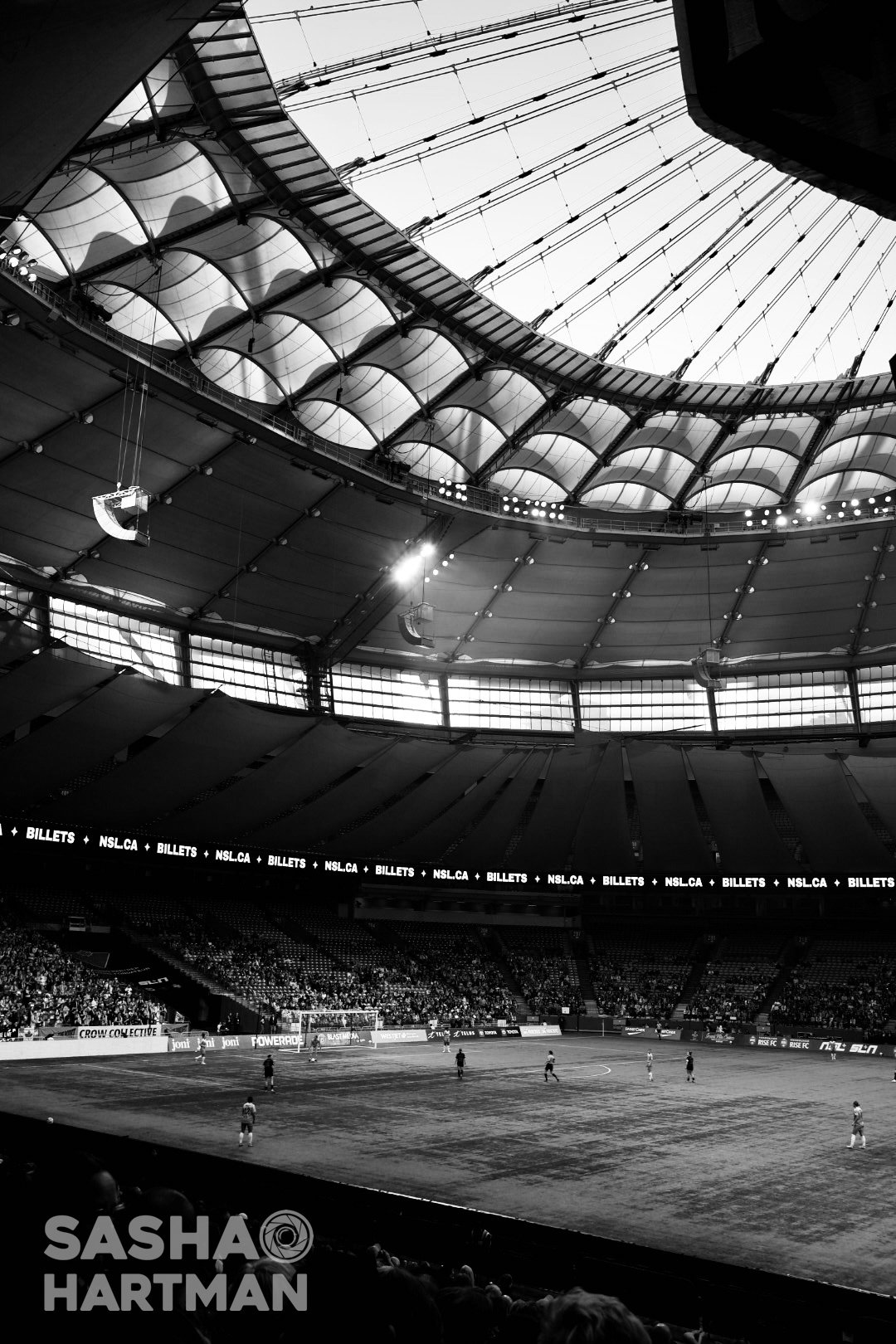 Interior of a large stadium with a curved, partially transparent roof. A soccer game is in progress on the field, with players scattered across the pitch. Audience seats are filled, and advertising banners are visible along the edges of the field.
