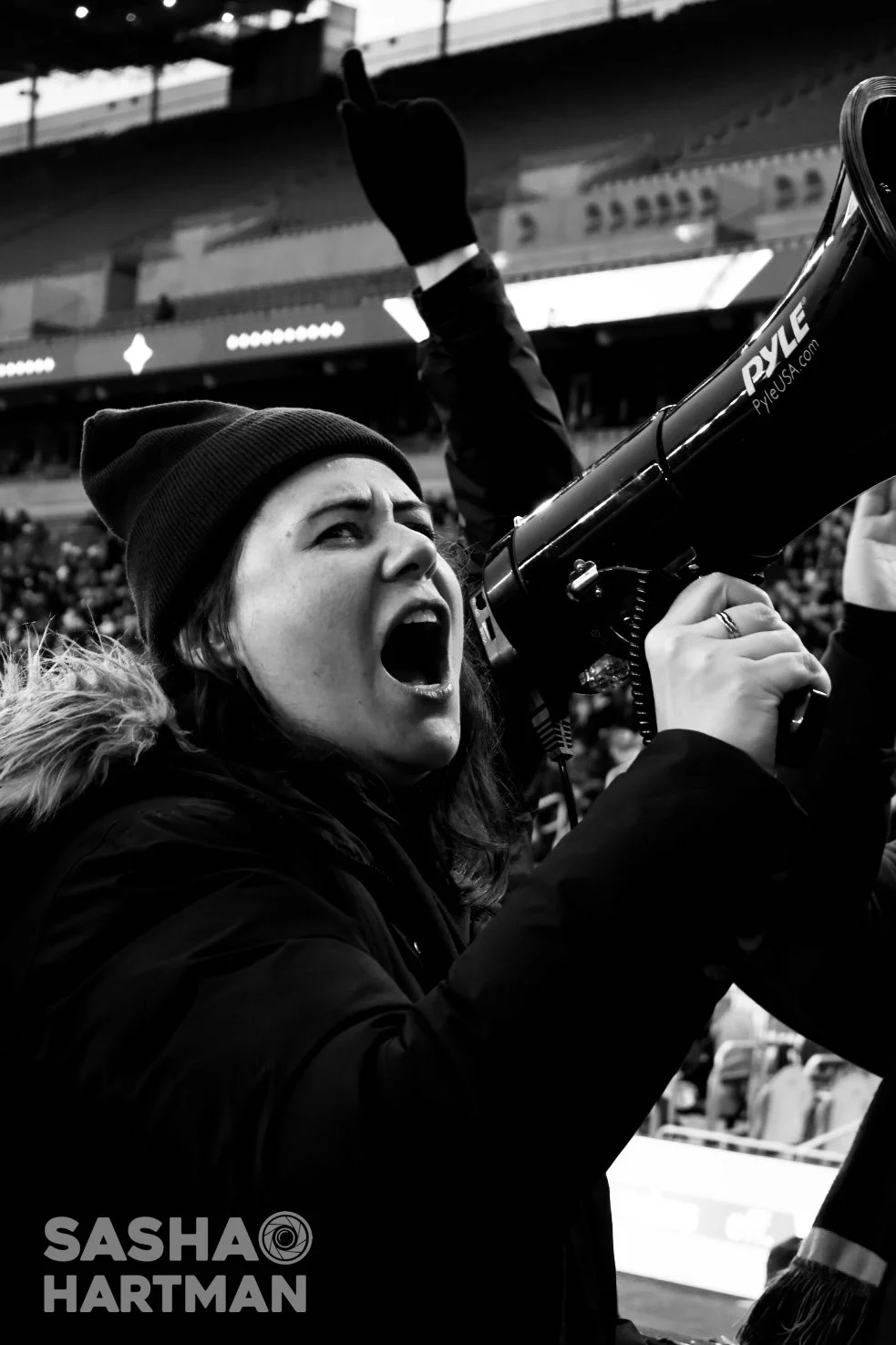 A woman wearing a beanie and winter coat shouting into a megaphone during a sports event in a stadium, with a crowd in the background.