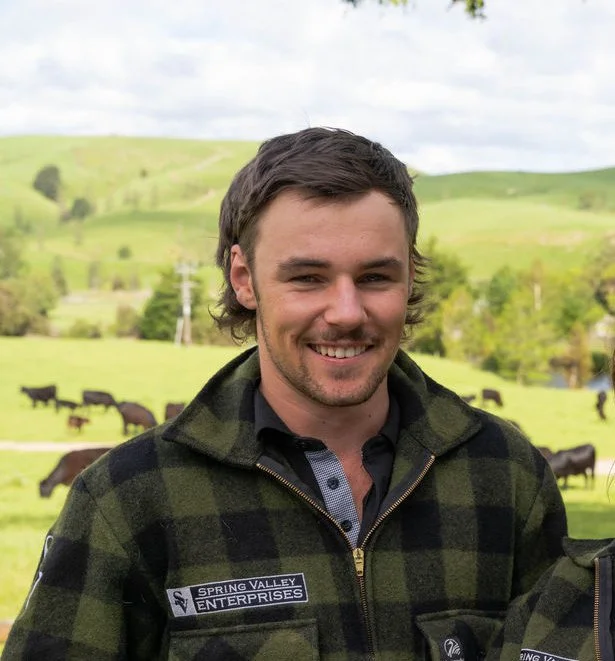 A man wearing a helmet and plaid jacket sitting on a red ATV, holding a large dog with a scenic green hillside and blue sky with clouds in the background.