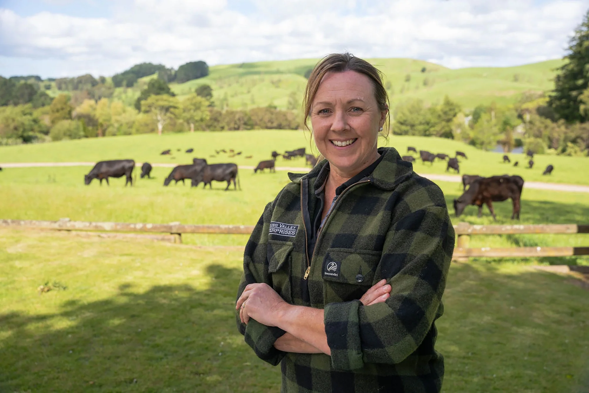 A woman in a plaid jacket standing with arms crossed outdoors with cows grazing in a green pasture behind her.