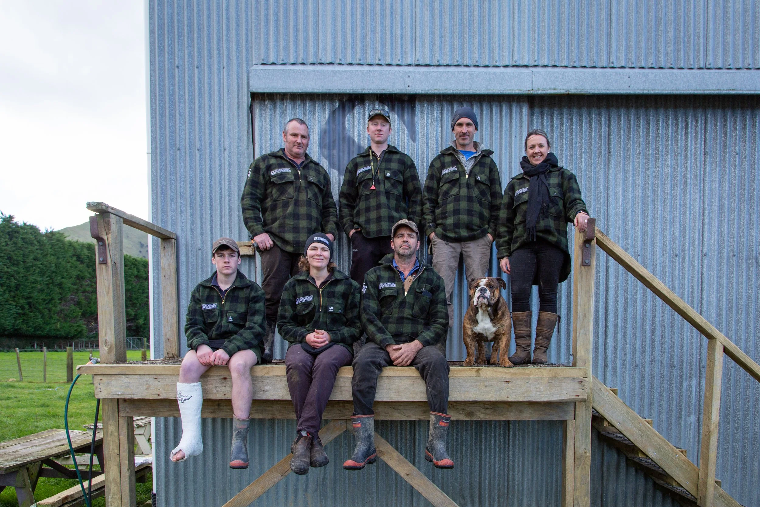 A group of seven people and one bulldog posing on a wooden platform in front of a metal building, all wearing matching dark green and black plaid jackets.