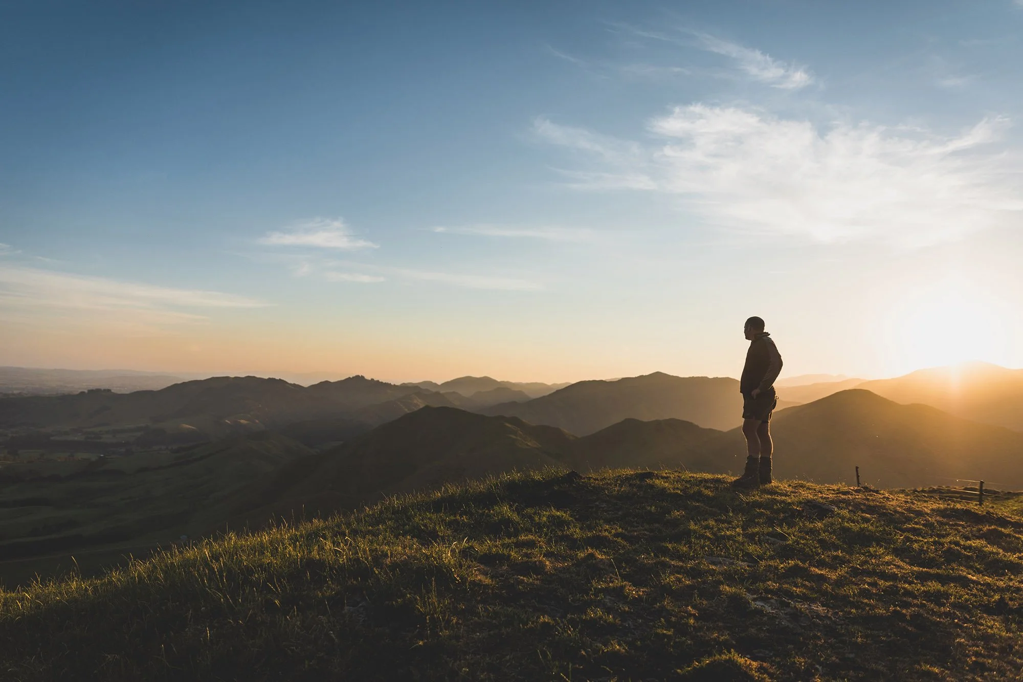 Person standing on a grassy hill at sunset, overlooking rolling hills and mountains.