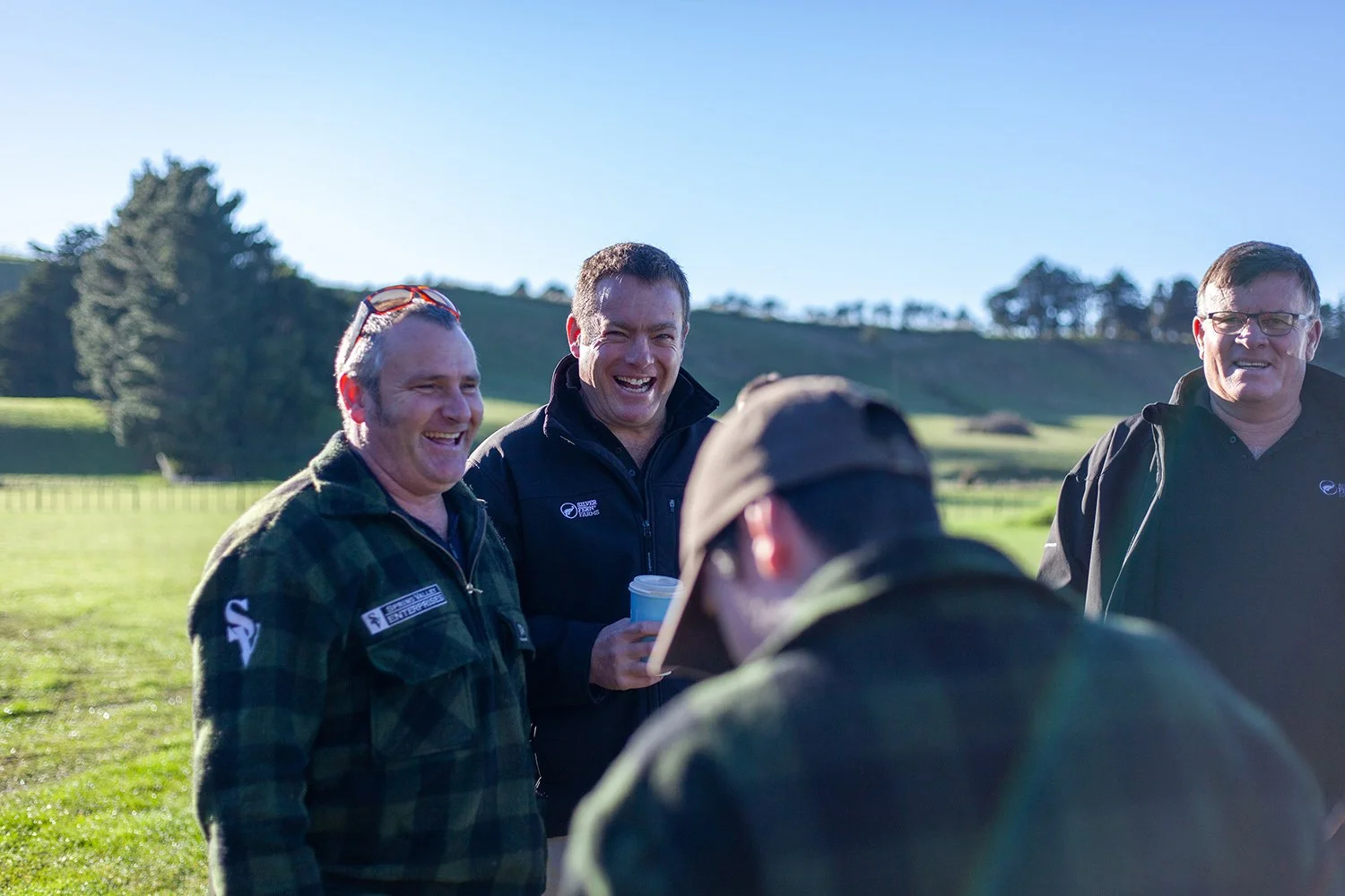 Four men outdoors on a sunny day, smiling and talking, with a grassy field and hills in the background.