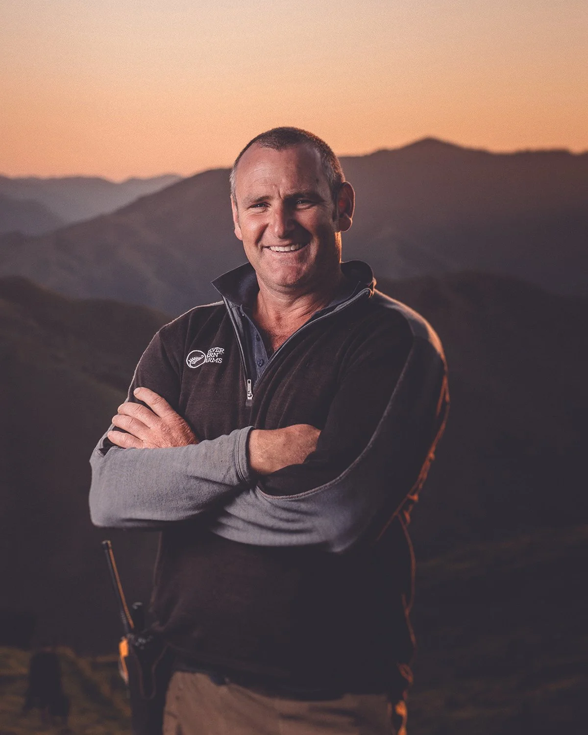 A smiling man standing with crossed arms outdoors during sunset, with mountains in the background.