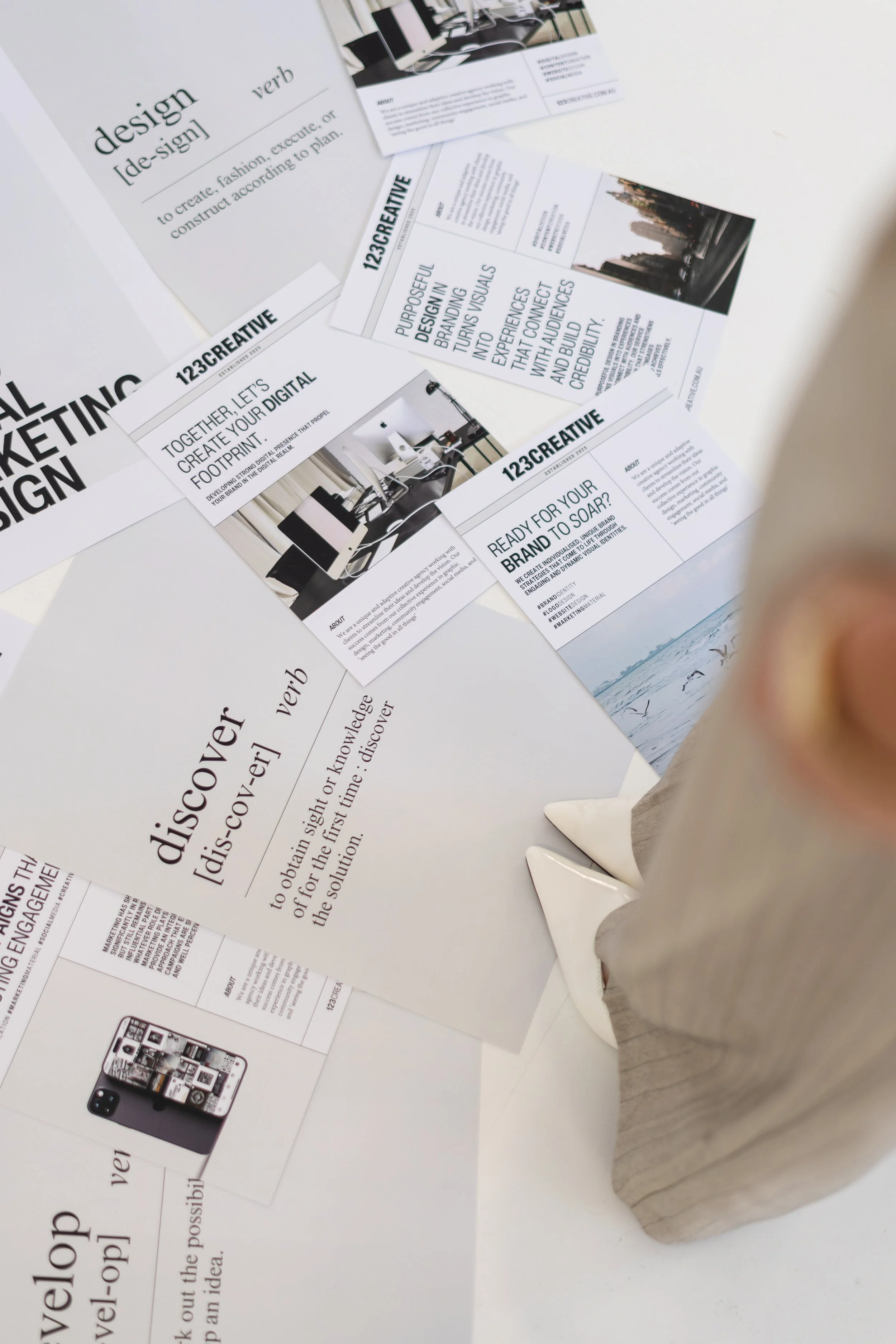 A person in beige high heels and beige pants standing over various printed marketing materials and brochures on a white surface.