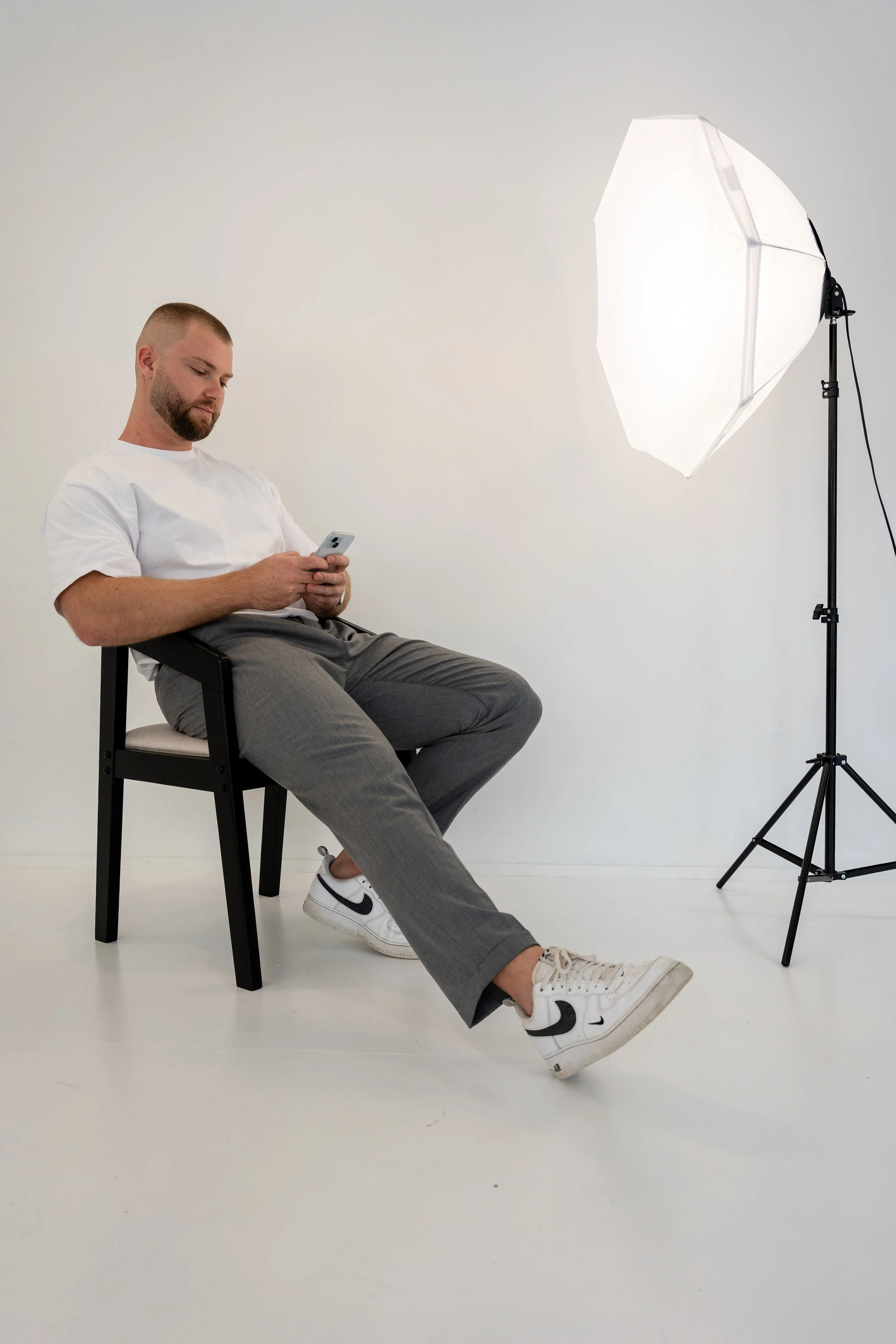 Man sitting on a black chair in a photography studio, using a smartphone, with a professional lighting setup nearby.