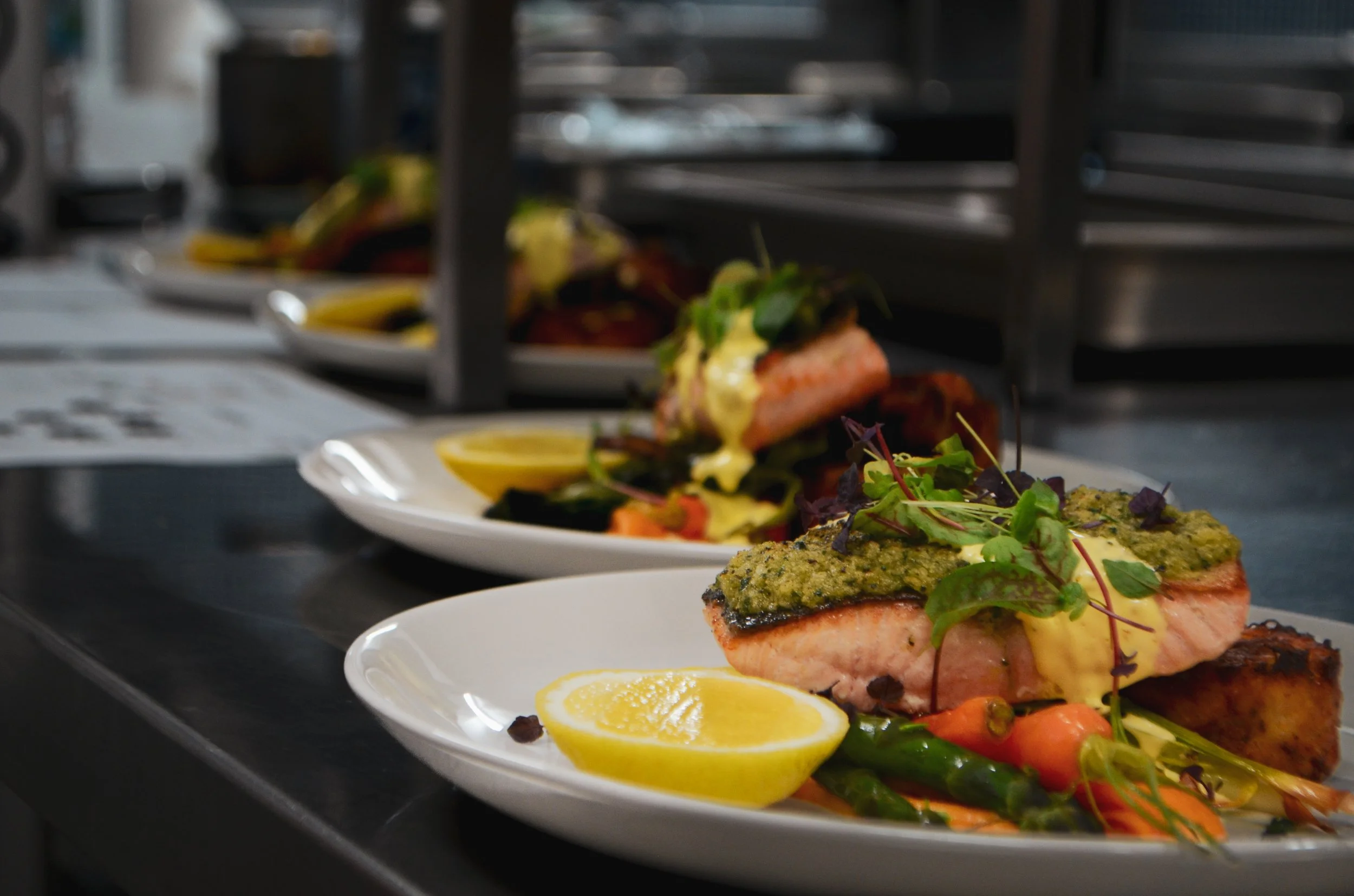 Close-up of prepared plates with salmon topped with herb crust, served with lemon wedges, cherry tomatoes, green beans, and microgreens in a professional kitchen setting.