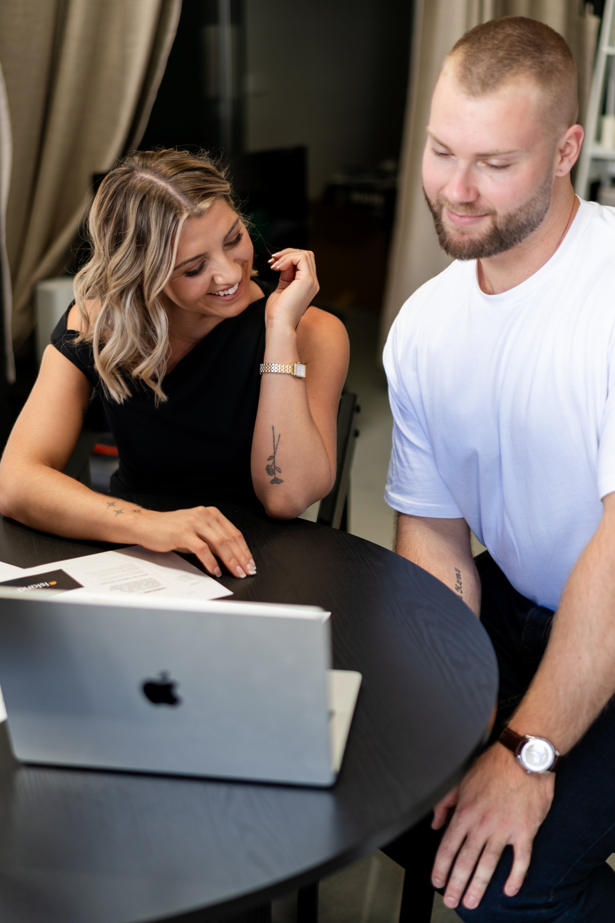 A woman and man sitting at a table, looking at a laptop. The woman is smiling, resting her head on her hand. The man is leaning forward, wearing a white T-shirt. There are papers on the table.